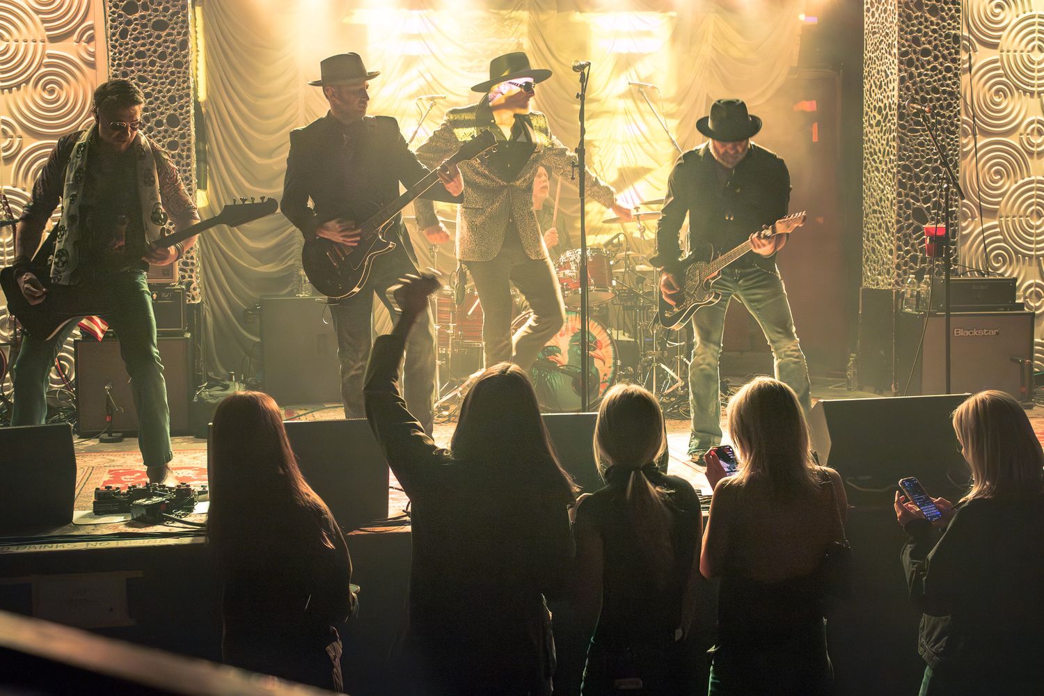 Crowd cheering with hands raised at a live rock band performing on a warmly lit indoor stage in Charlotte, NC.