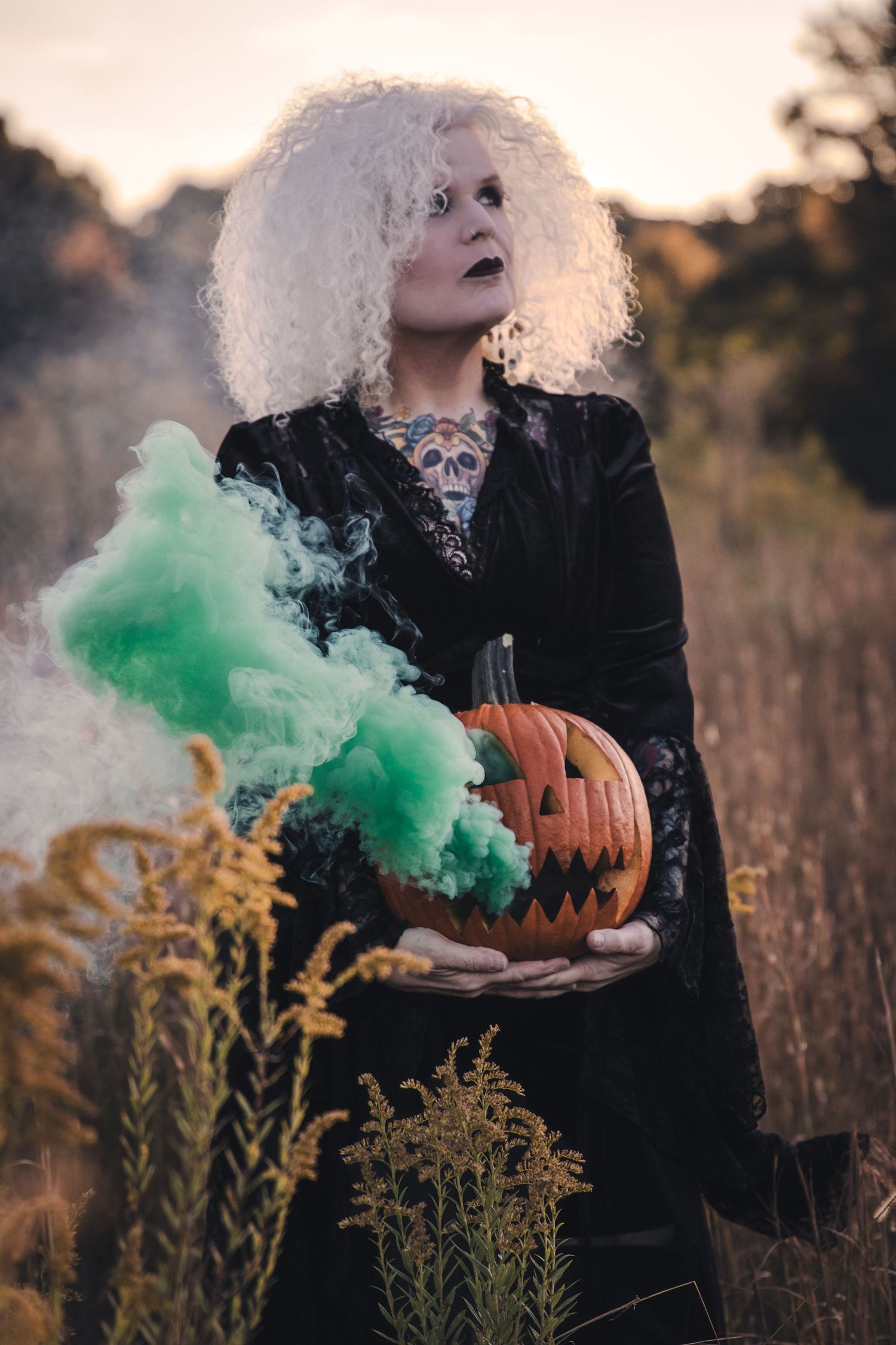 Creative portrait session with woman in witch costume holding a smoking jack-o-lantern pumpkin in an autumn field at sunset.