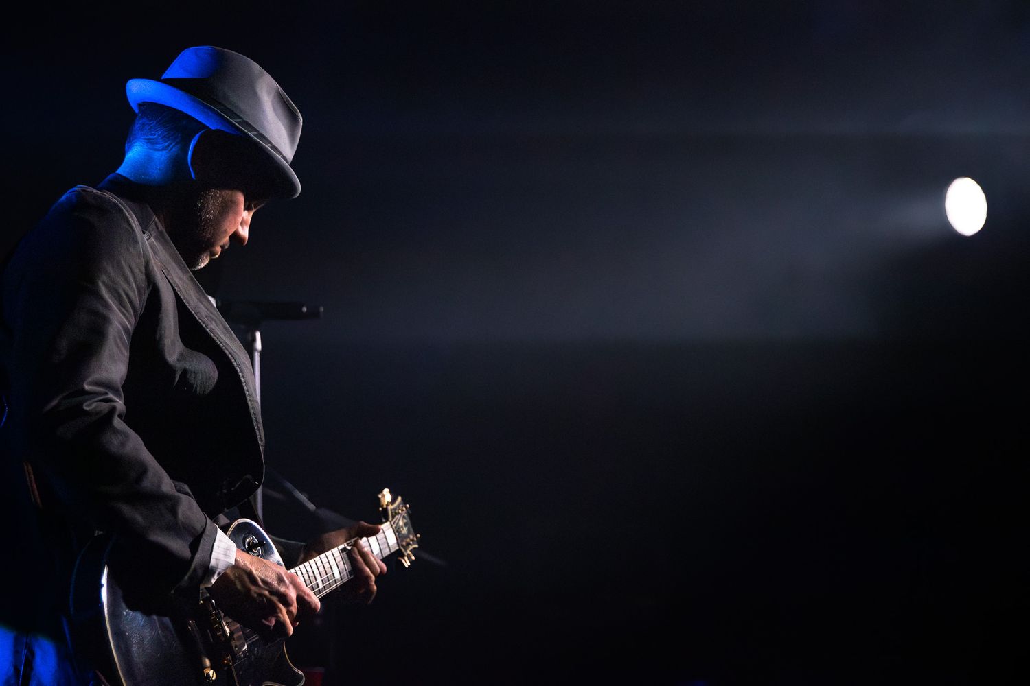 Guitarist in black suit and fedora performing on stage under dramatic blue and white spotlight in Charlotte, NC.