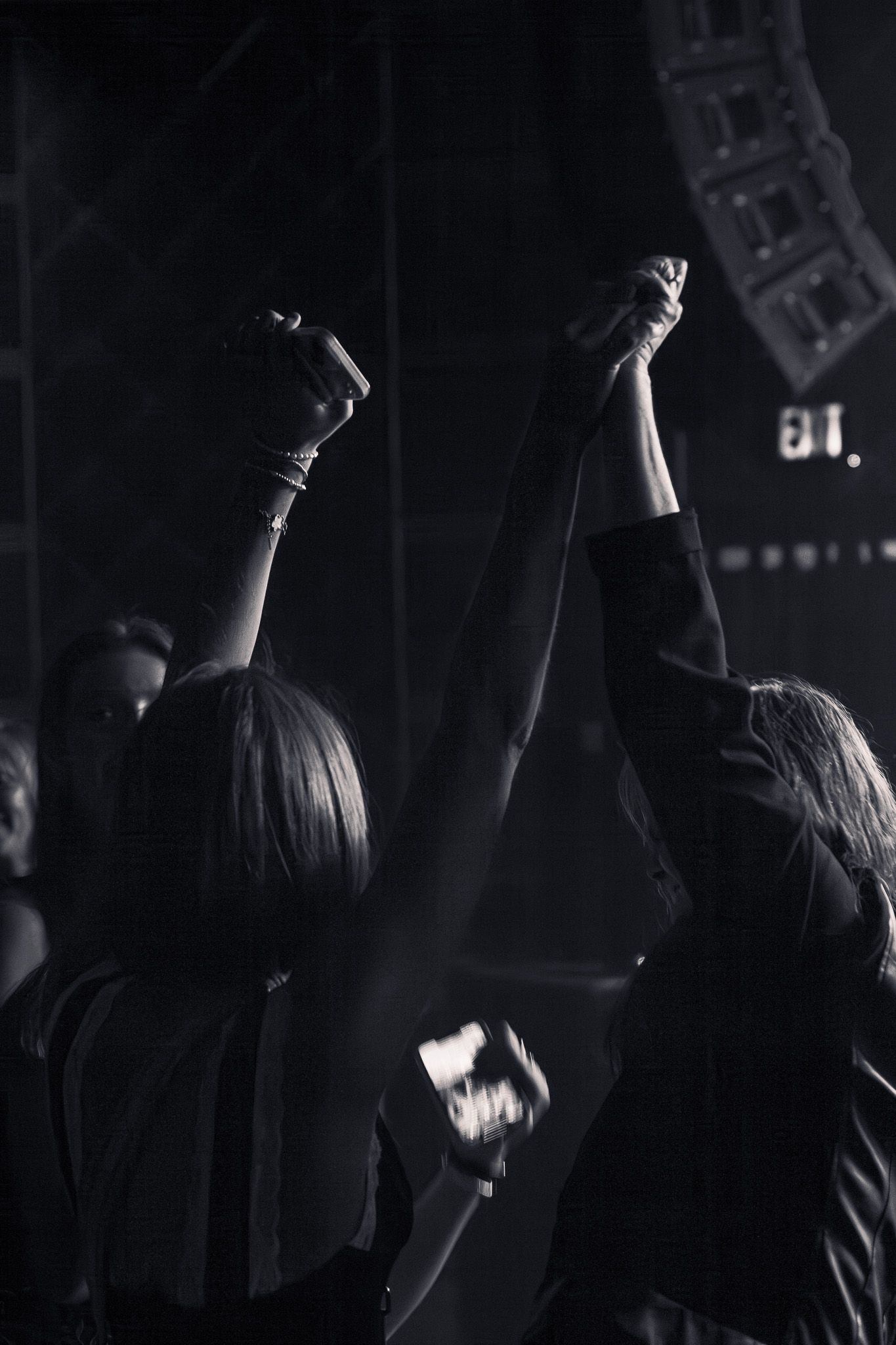 Black and white silhouette of two women raising hands together at a live concert.