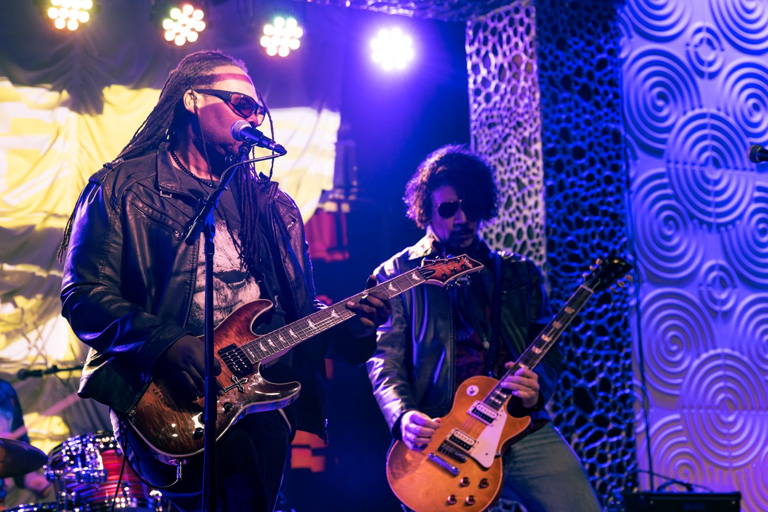 Two guitarists performing on stage with colorful purple lighting and patterned backdrop.