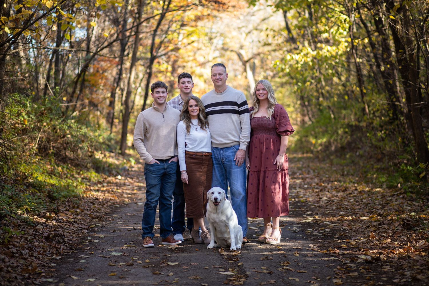 Happy family of four with a white dog posing on a tree-lined autumn path covered in fallen leaves.