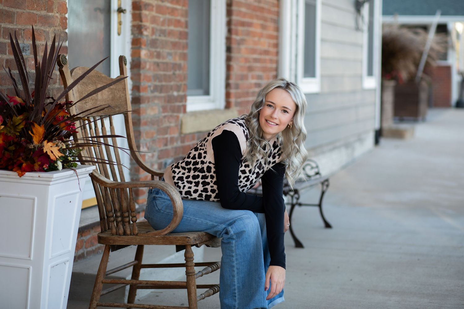 Woman with gray hair sits smiling on a wooden chair on a brick building porch with fall floral decor.