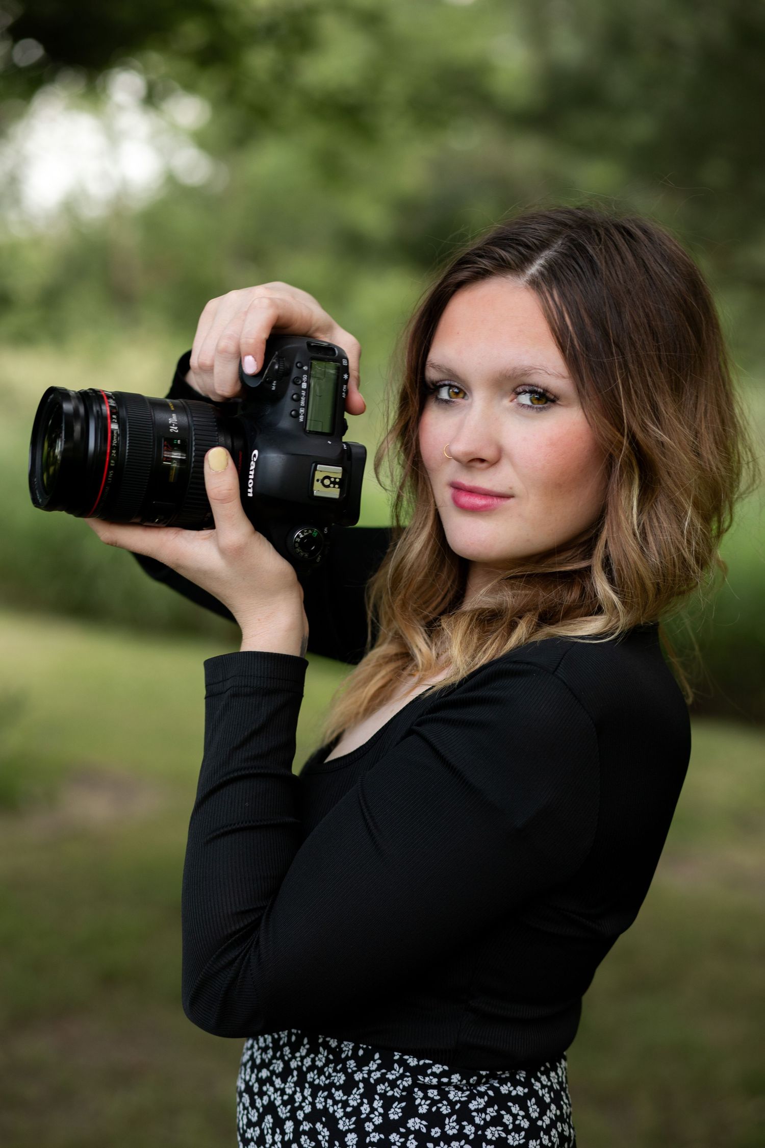Young woman photographer holding a DSLR camera outdoors in a park, wearing a black top and floral skirt.