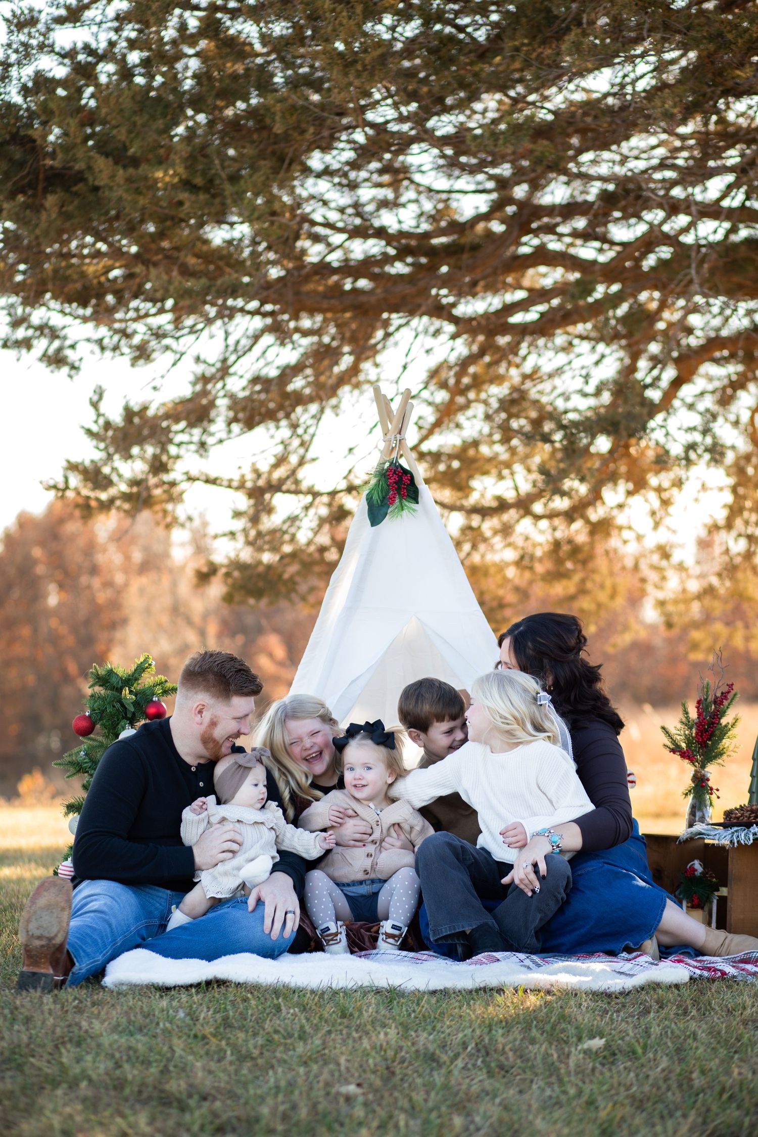 Multi-generational family gathered on a blanket near a teepee at golden hour in an autumn park.