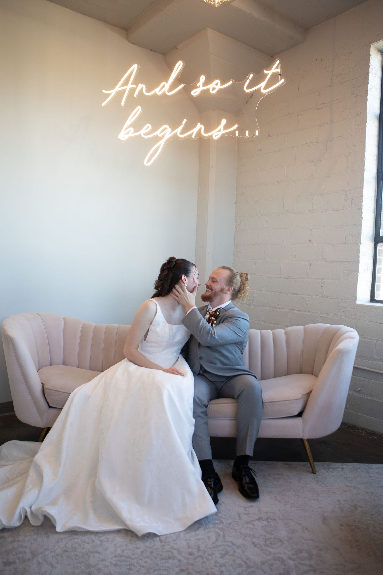 Bride and groom sitting on a pink sofa beneath a glowing neon sign reading And so it begins.