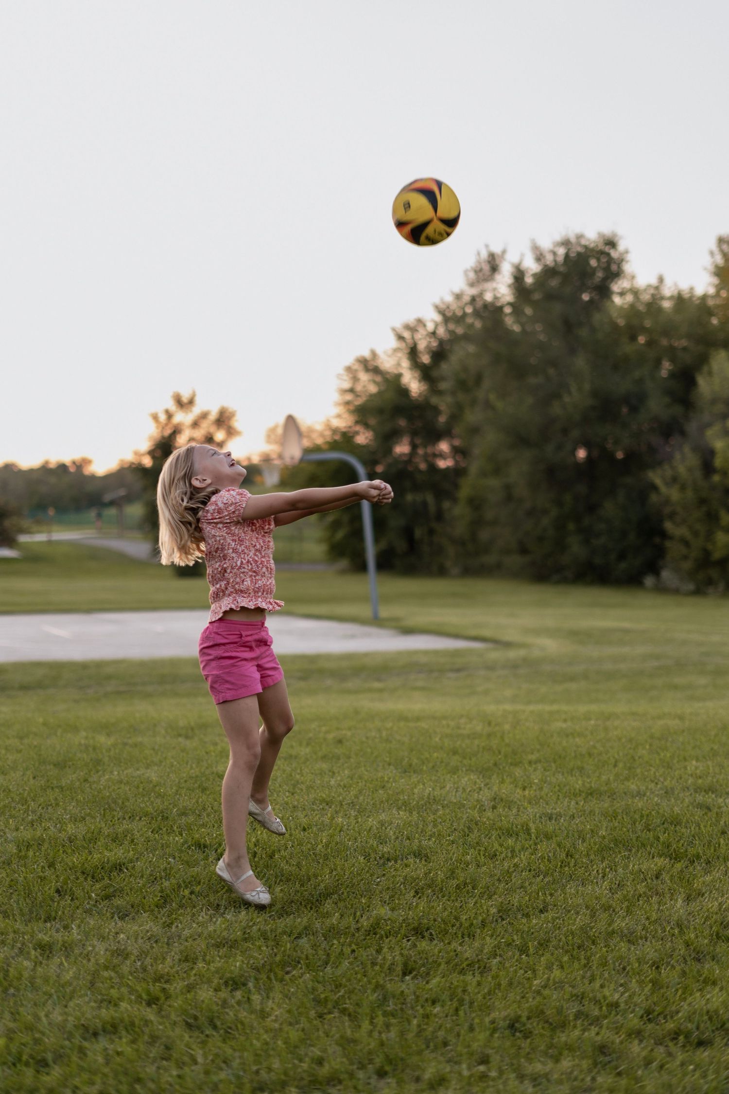 Young girl playing volleyball outside on grassy field at sunset in pink summer outfit.