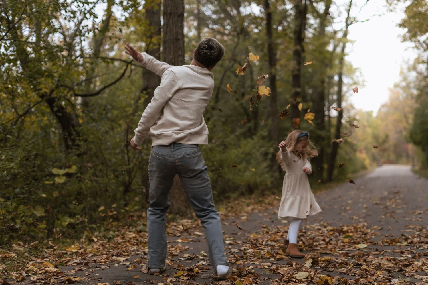 Siblings throwing leaves together on an autumn path surrounded by fallen leaves and forest trees.