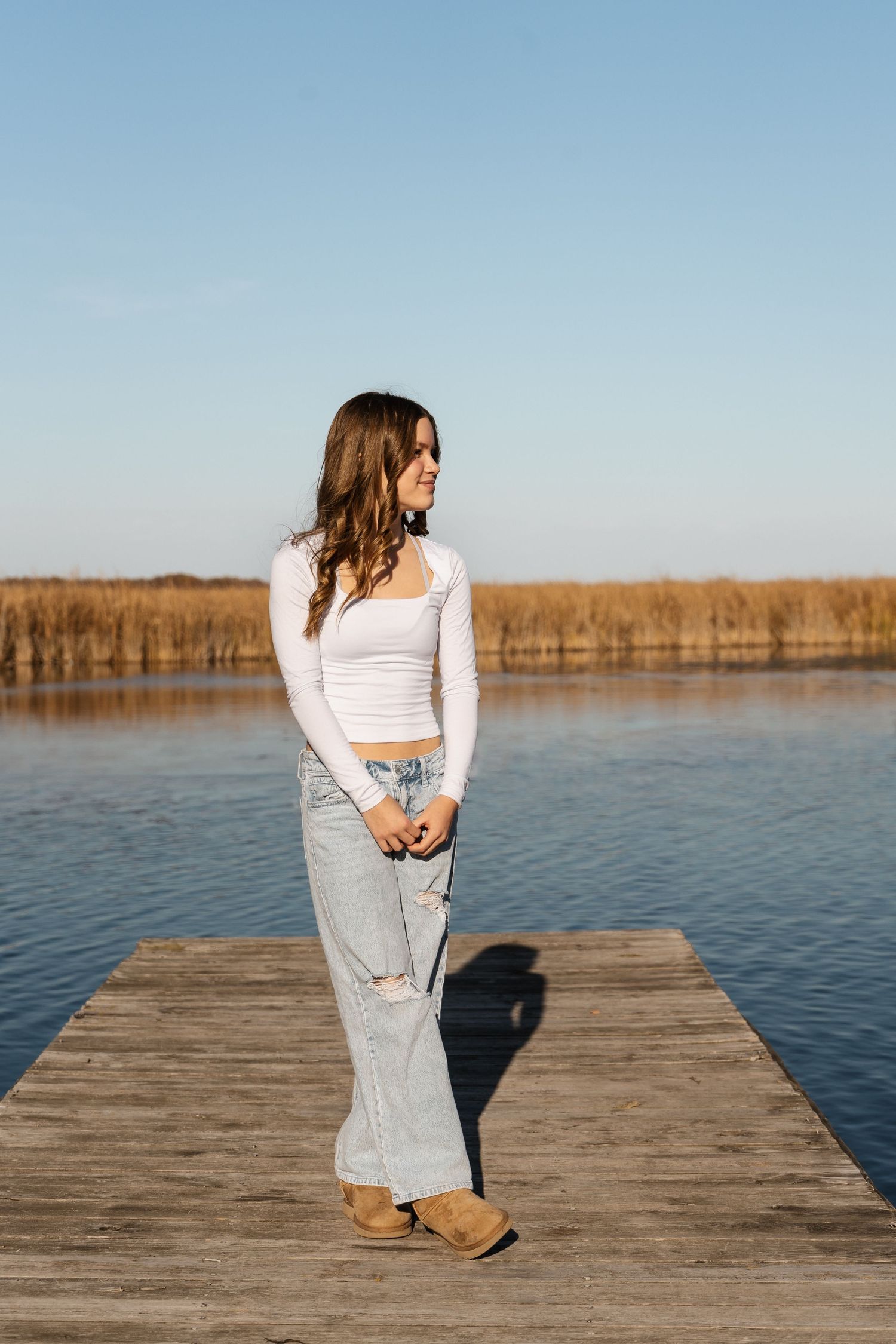 A person in a white top and jeans stands on a wooden dock overlooking a tranquil lake.
