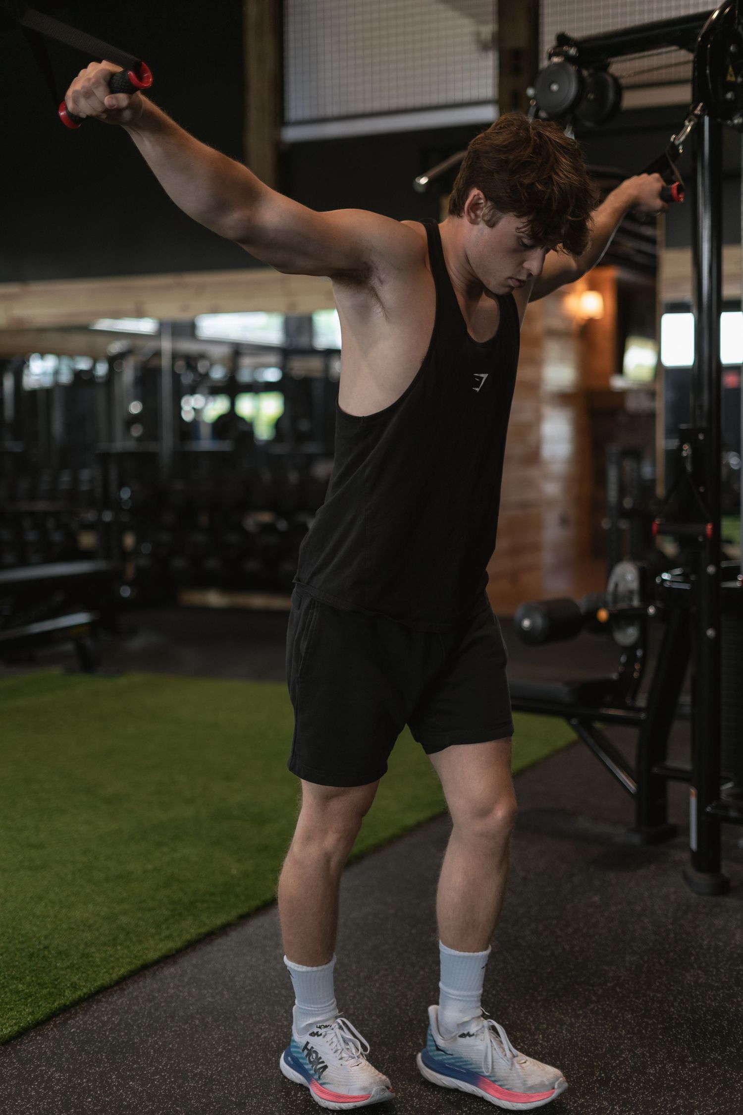 Person in black tank top and shorts doing lateral cable exercises in modern gym setting.