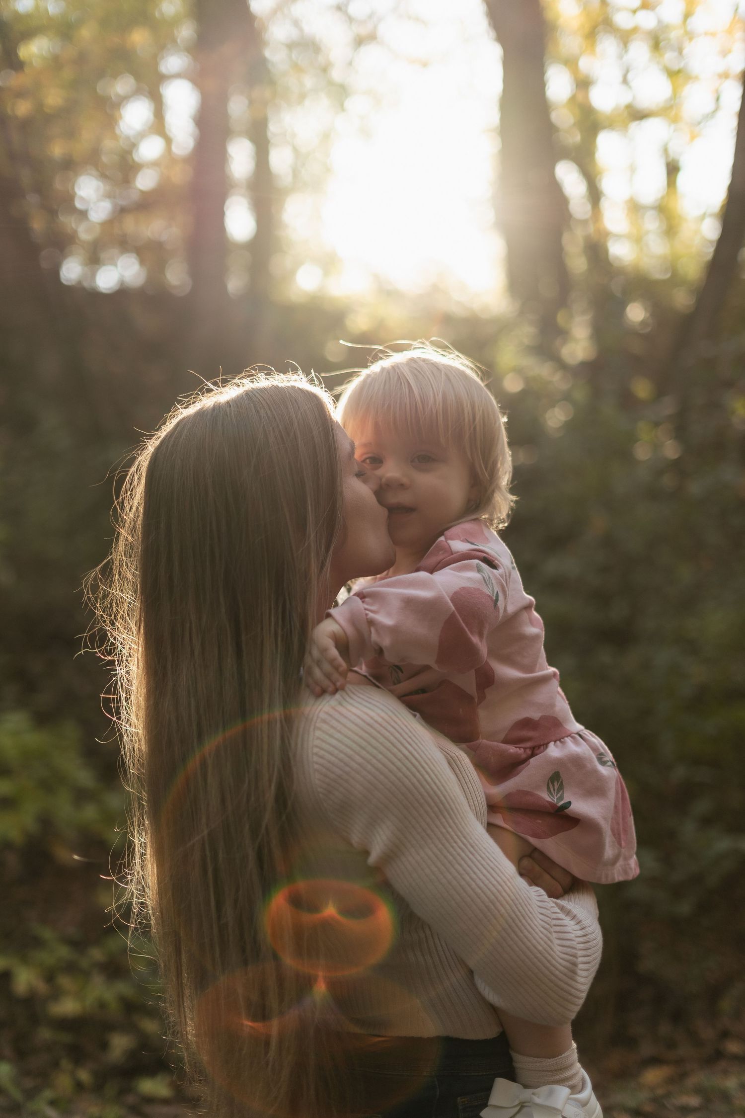 Tender outdoor moment between parent and child backlit by golden sunlight through trees.