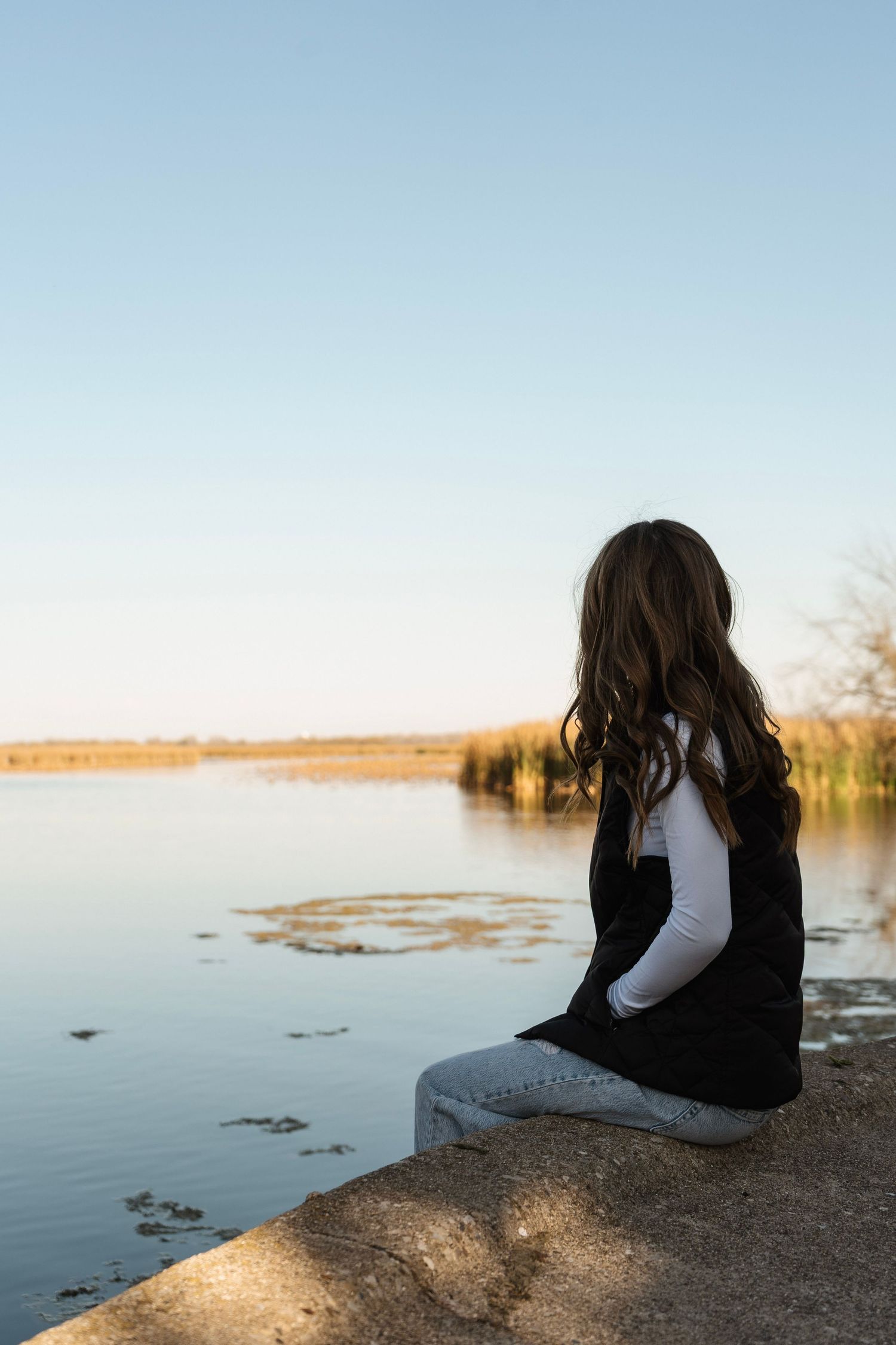 Girl sitting by tranquil lake during sunset, surrounded by autumn reeds and calm waters.