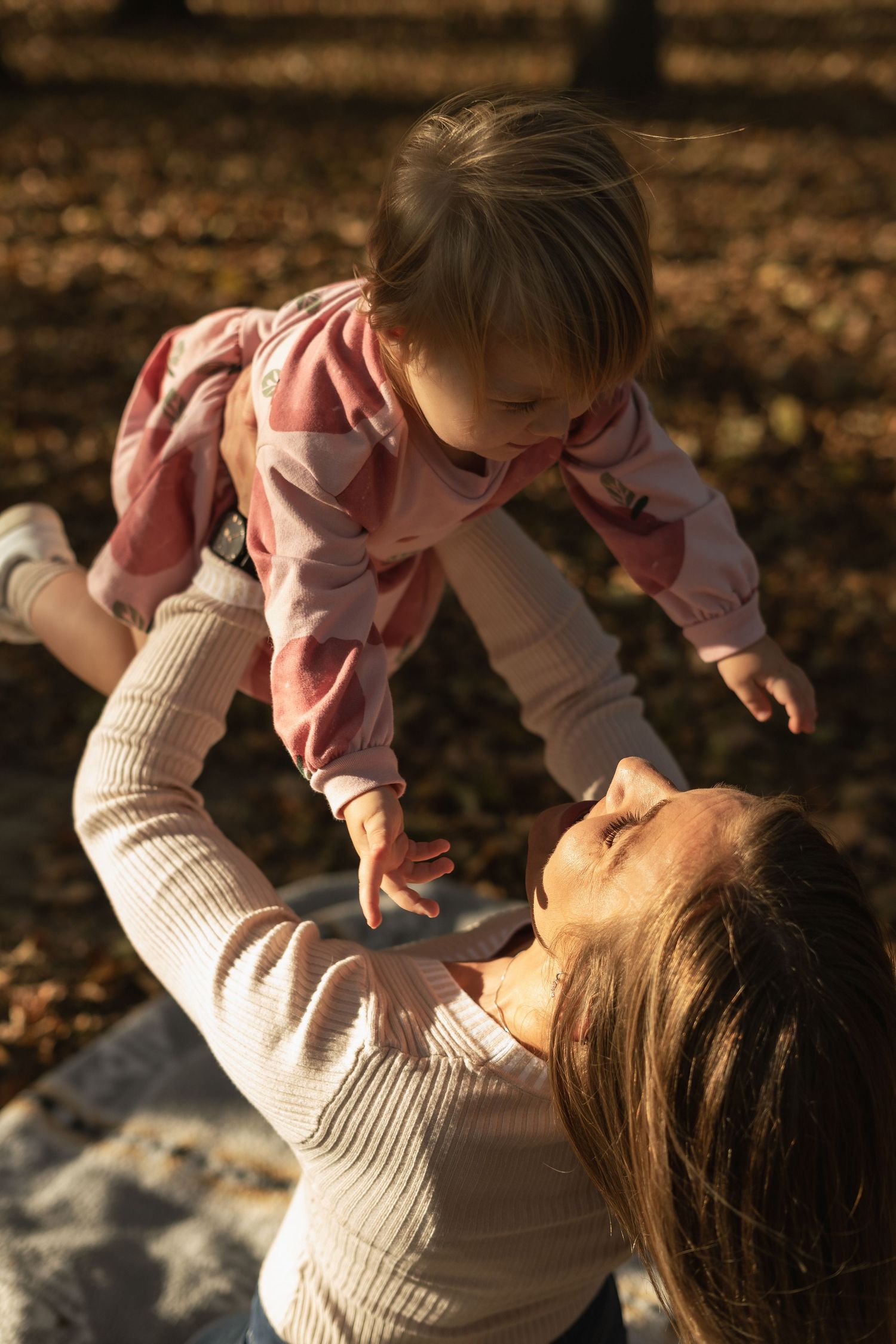 A tender moment captured as a caregiver lifts and playfully interacts with a small child against an autumn backdrop.