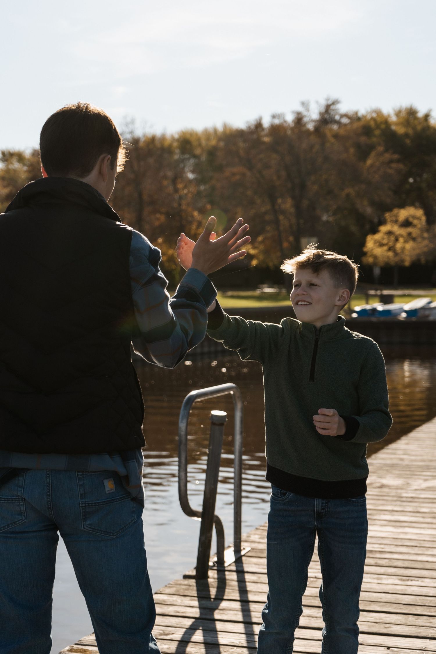 Cousins share an energetic moment while standing on a wooden dock by a lake during autumn.