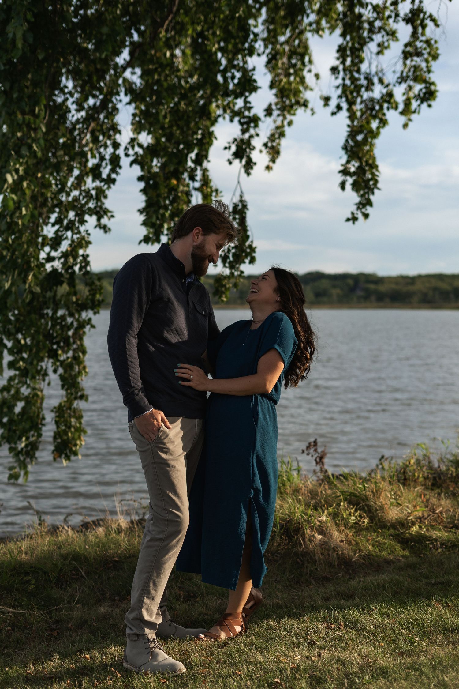 A romantic couple embraces under hanging tree branches beside a scenic lake on a calm summer evening.