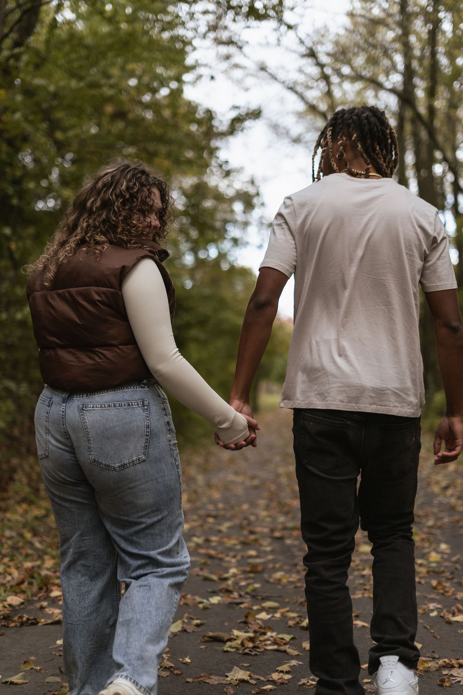 Couple holding hands while walking on a leaf-covered path through autumn woods.