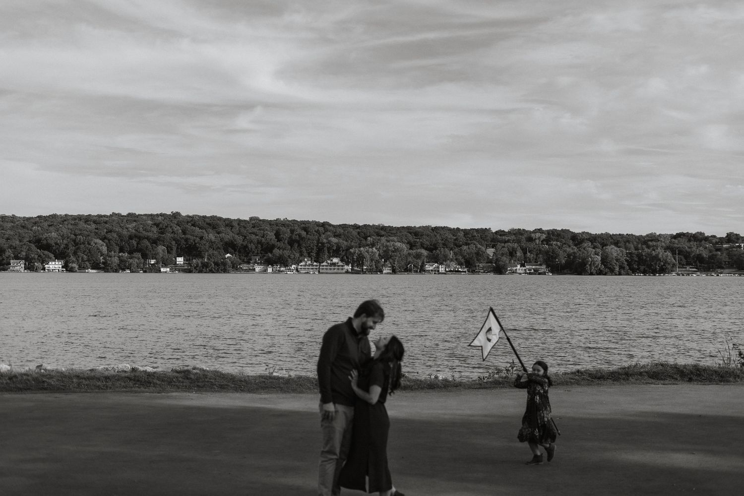 A black and white photo of a couple embraces by a lake while a small child plays with a balloon nearby.