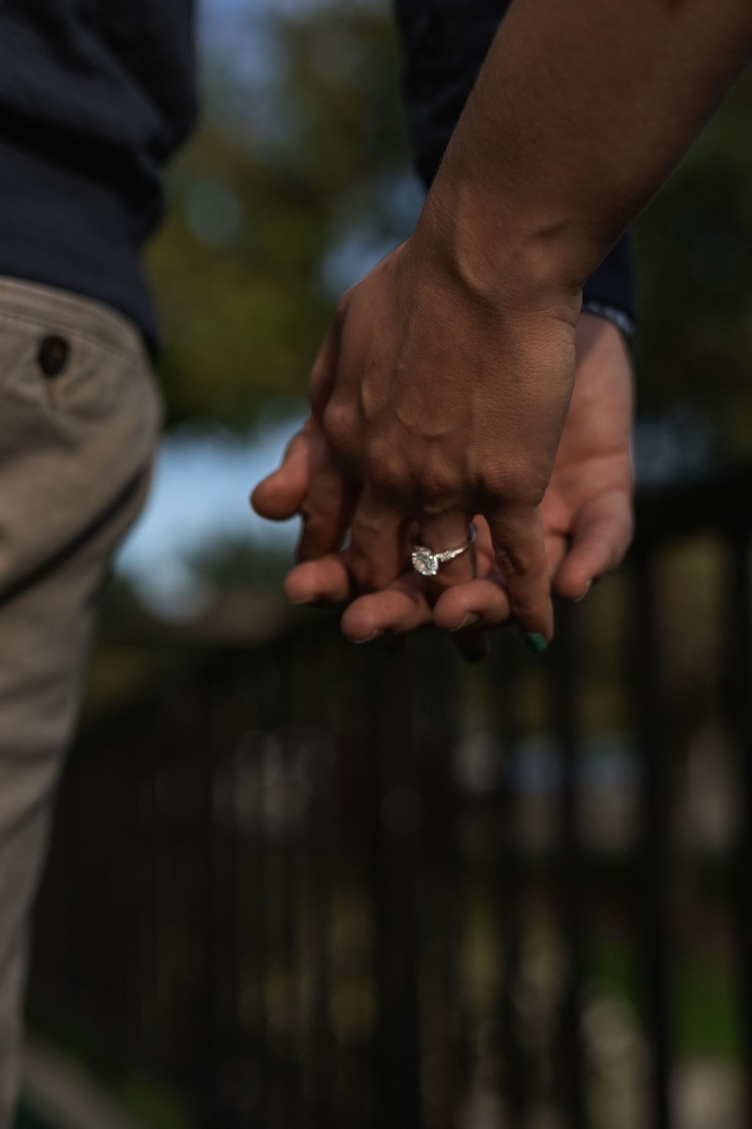 Close up photo series of hands passing a diamond engagement ring between them against a blurred outdoor background.