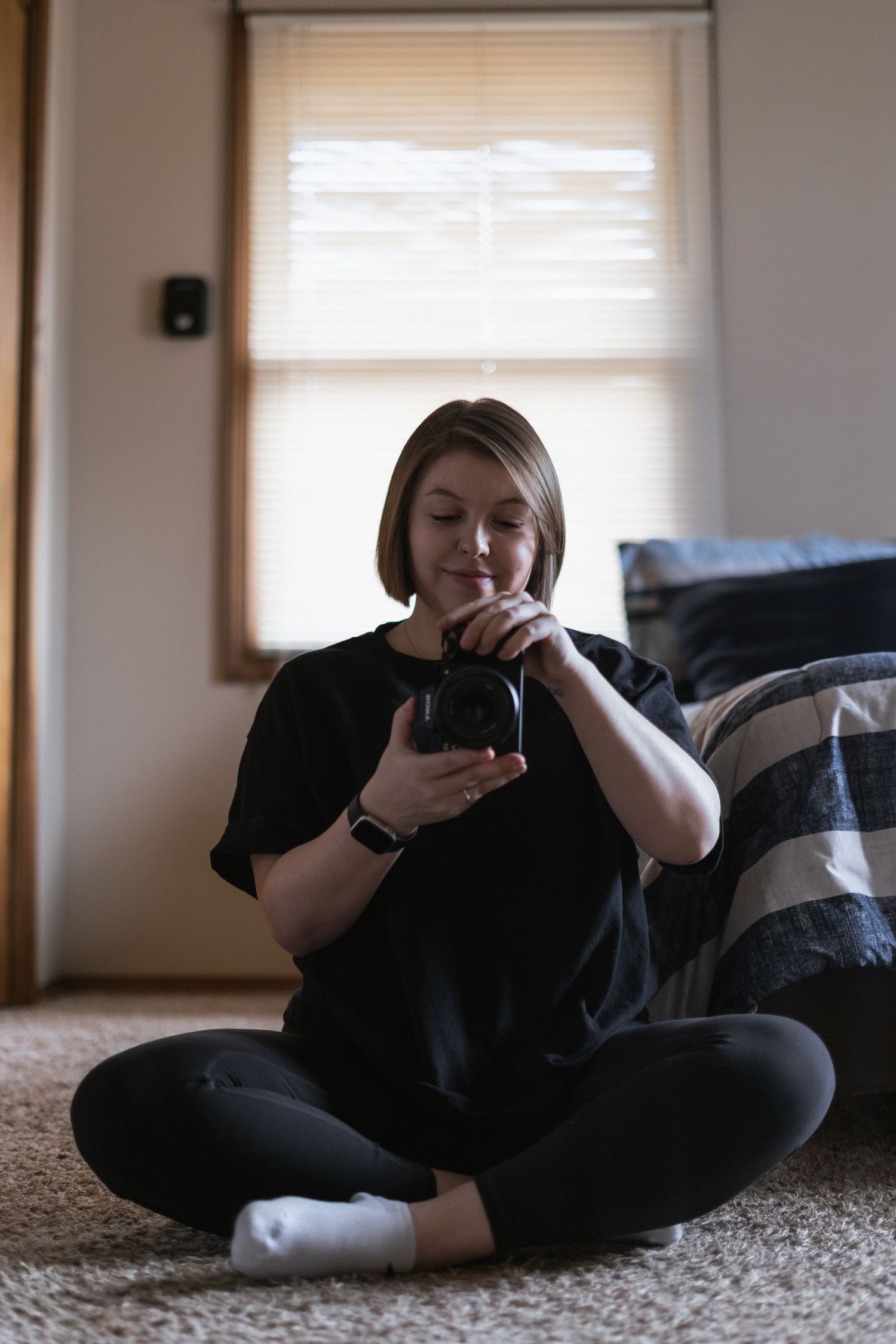 Photographer on carpet in casual black clothes holds a camera near a window with natural lighting.