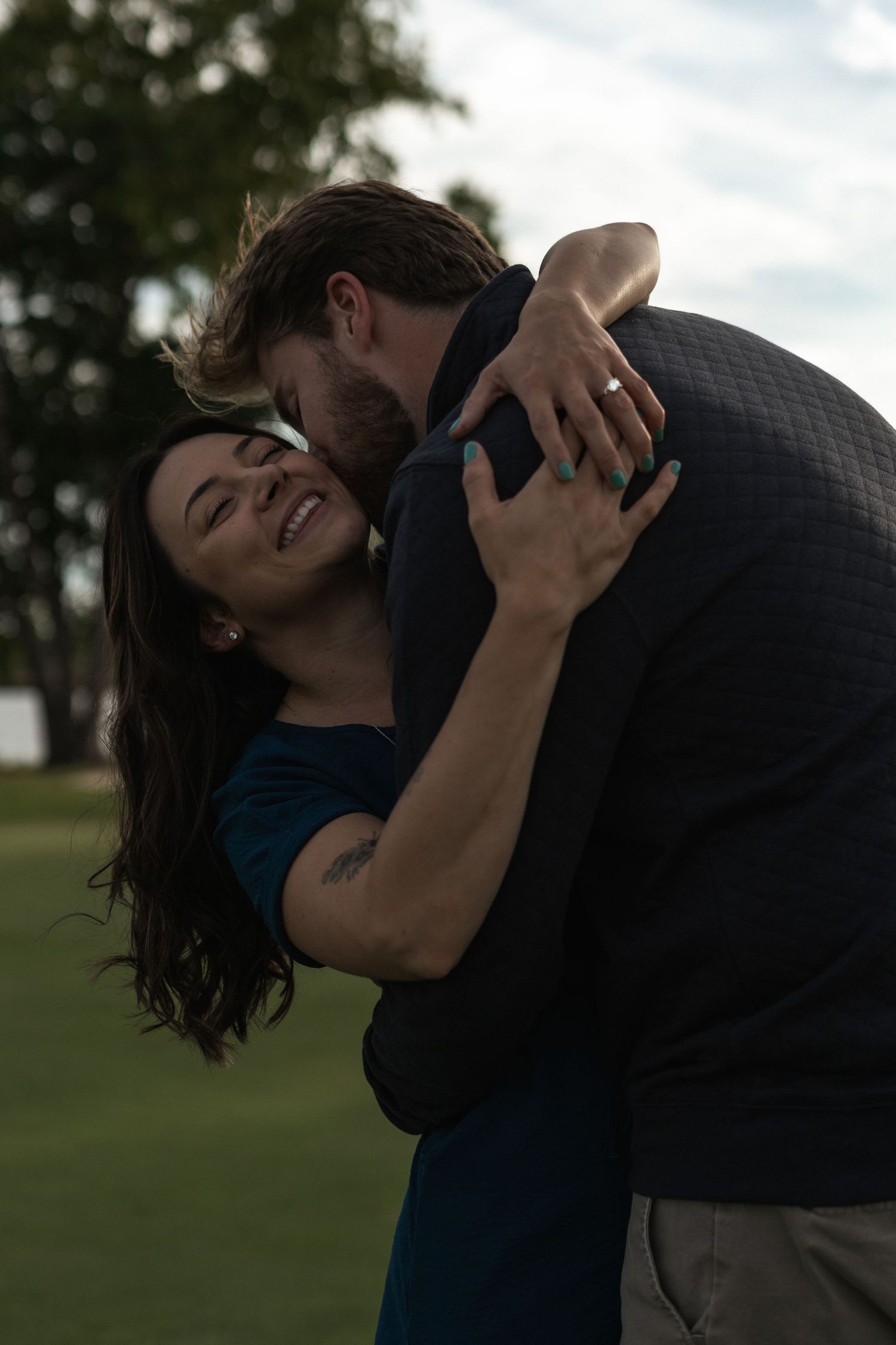 Couple embracing and laughing together outdoors on a grassy field during a romantic photoshoot at dusk.