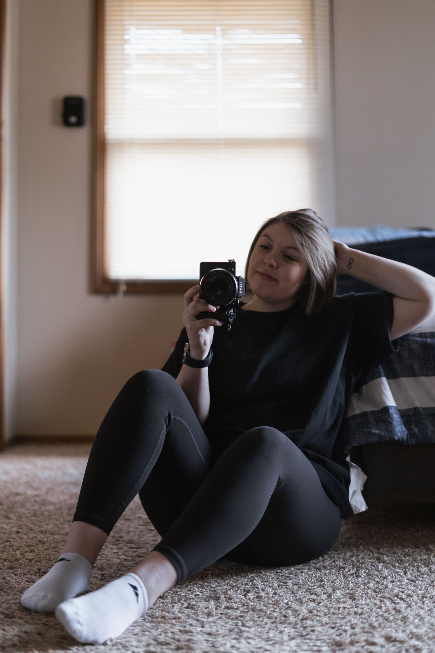 Photographer on carpet in casual black clothes holds a camera near a window with natural lighting.