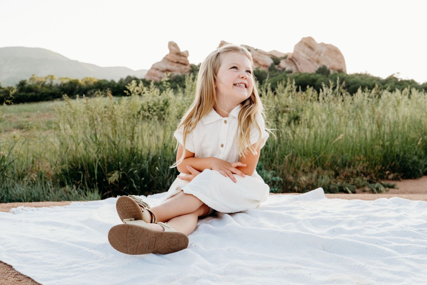 Young girl sitting on white blanket, legs crossed, daughter looking off into distance smiling, tall grasses behind her.