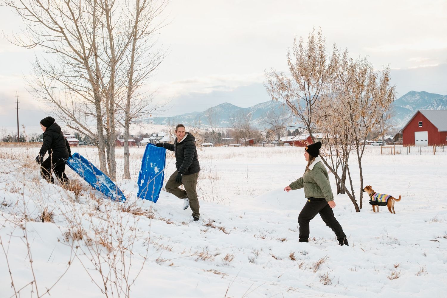 Son, father and mother walking up sledding hill on snowy day.  Trees, barn and Boulder Flat Irons in background.