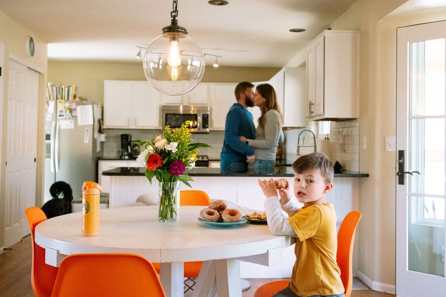 Boy sitting at the table eating a donut, parents cuddling in the background by the kitchen sink.