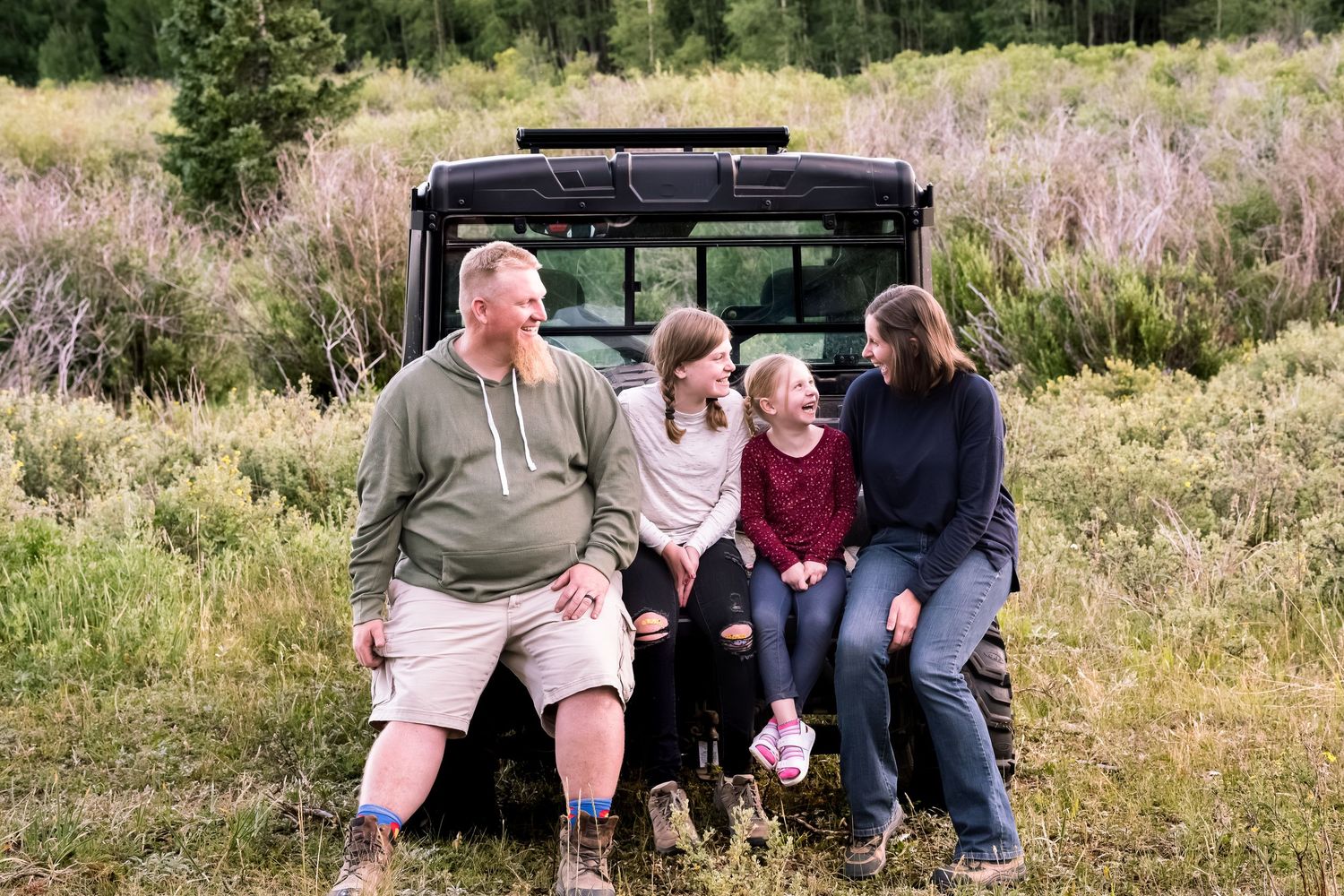 Father, two daughters and mother sitting on the back of a UTV in a green mountain meadow, all laughing.