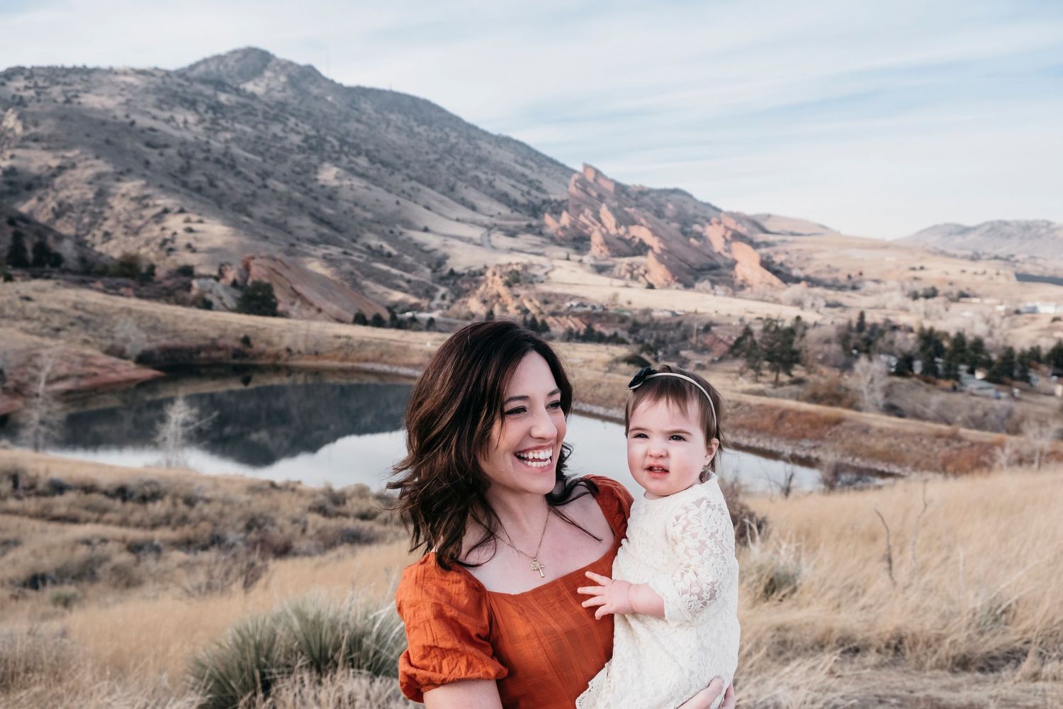 Mother standing, holding baby girl, pond and Red Rocks in the background.