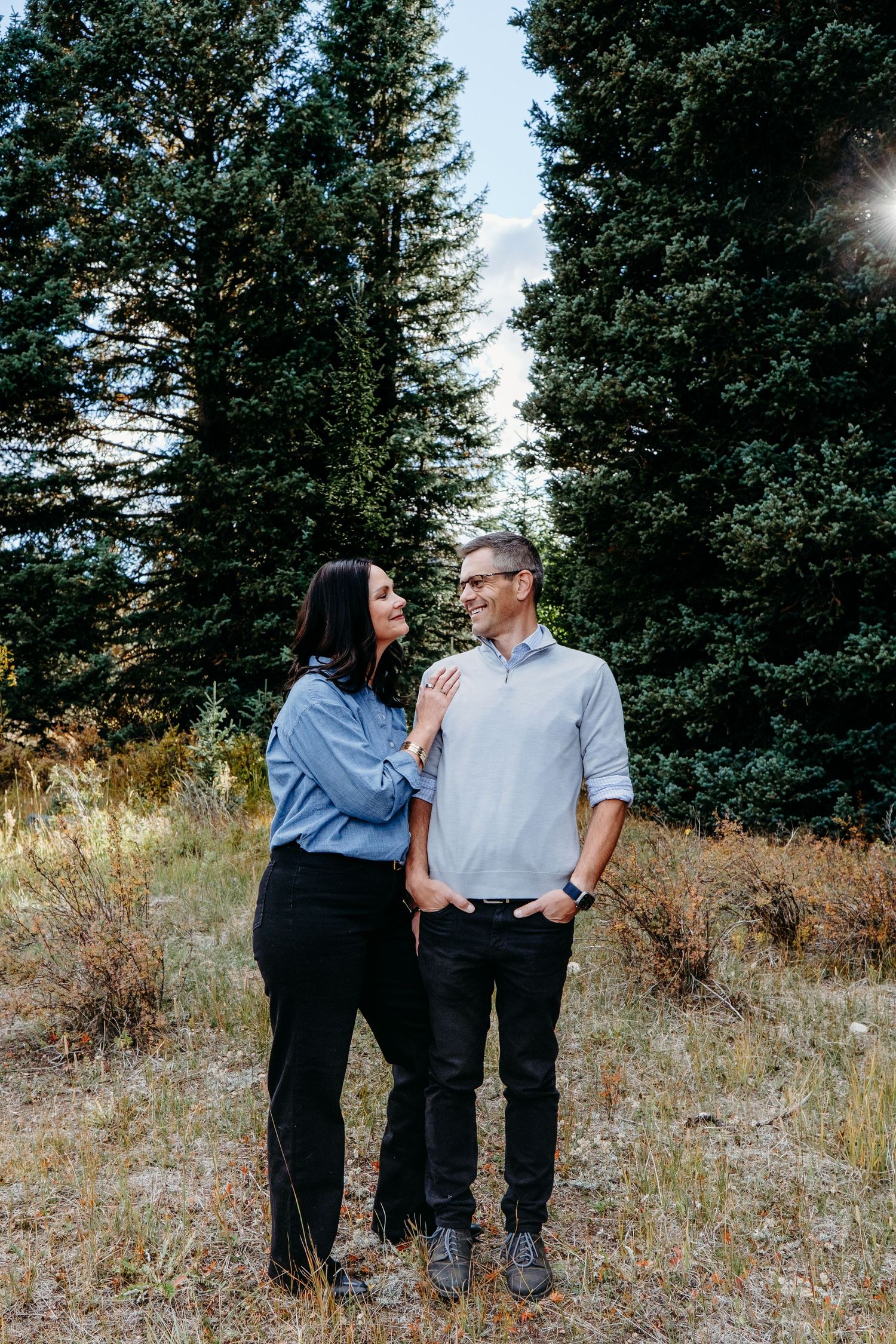 Husband and wife posing in front of towering evergreen trees in Colorado mountain session.