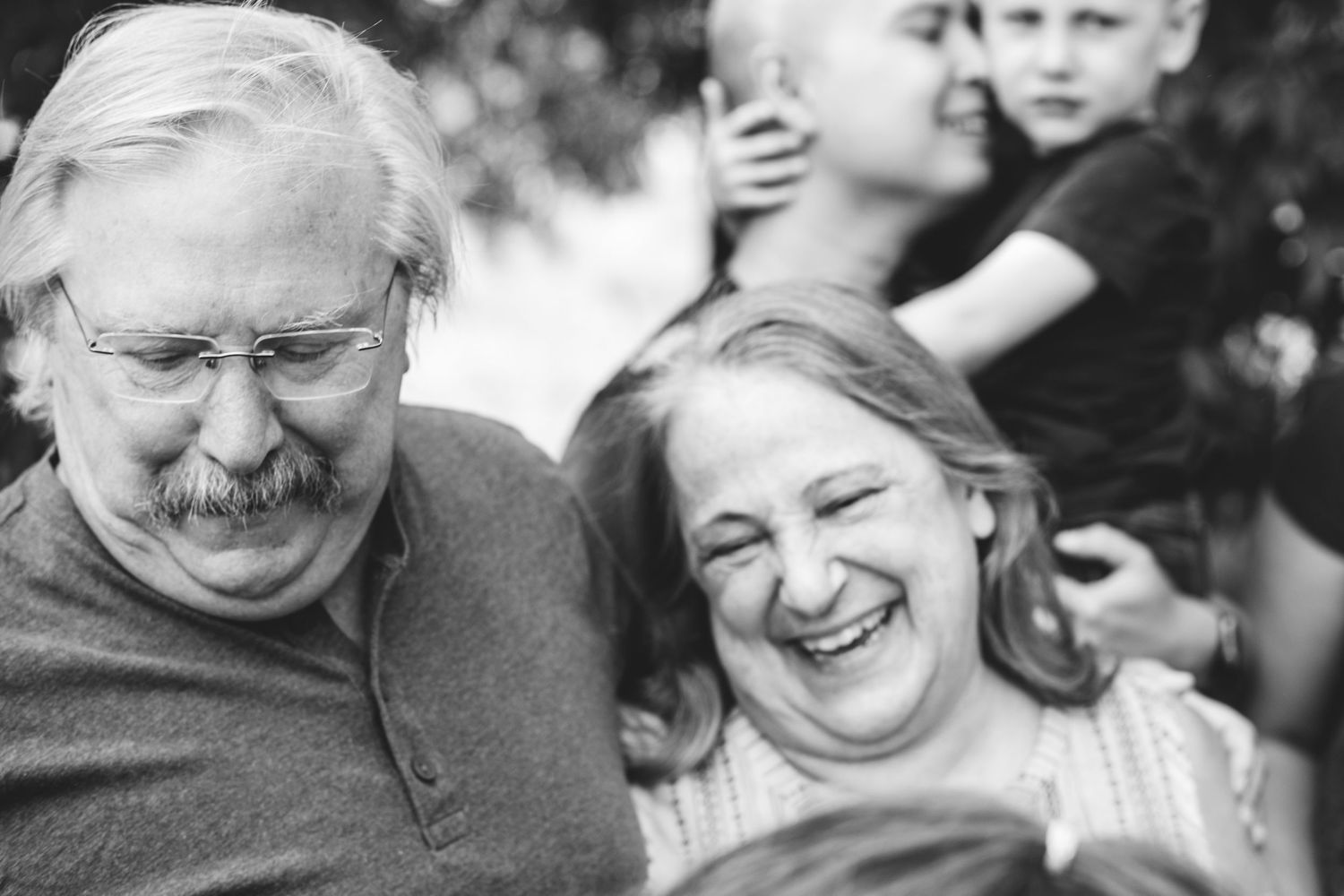Black and white layer portrait of grandfather and grandmother smiling, father holding son in background, snuggling.