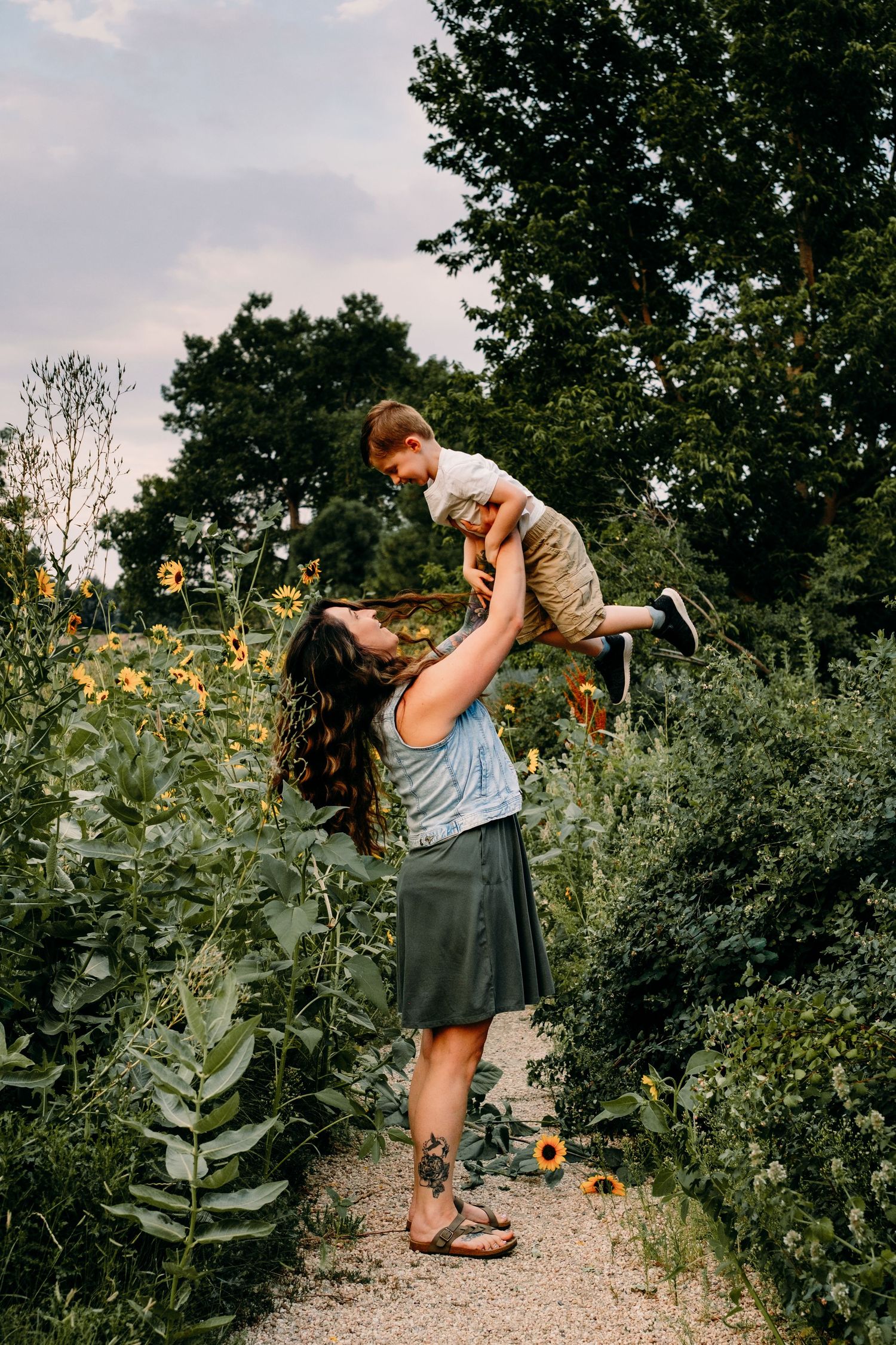 Playful documentary photography motherhood session, mother tossing toddler son into the air, both surrounded by sunflowers.