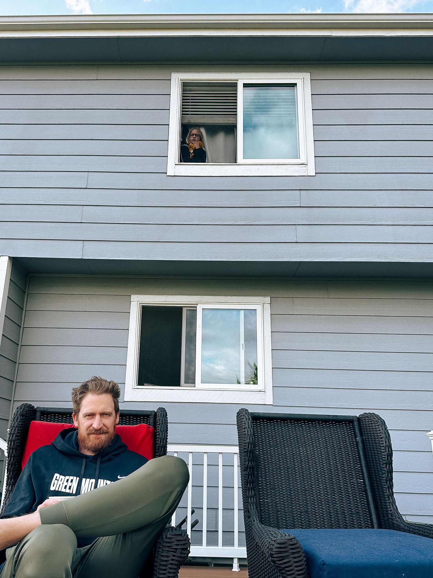 Father sitting on backyard deck, daughter in window of second story behind him.
