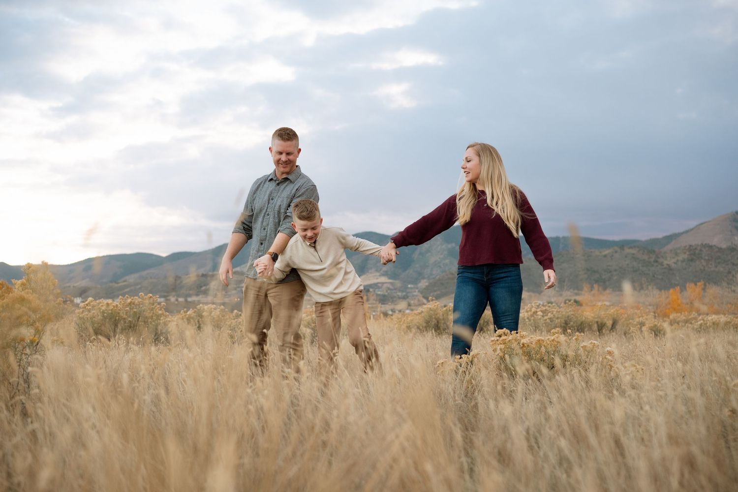 Playful family of three in tall grasses in Bear Creek Lake Park in Lakewood, foothills in the background.