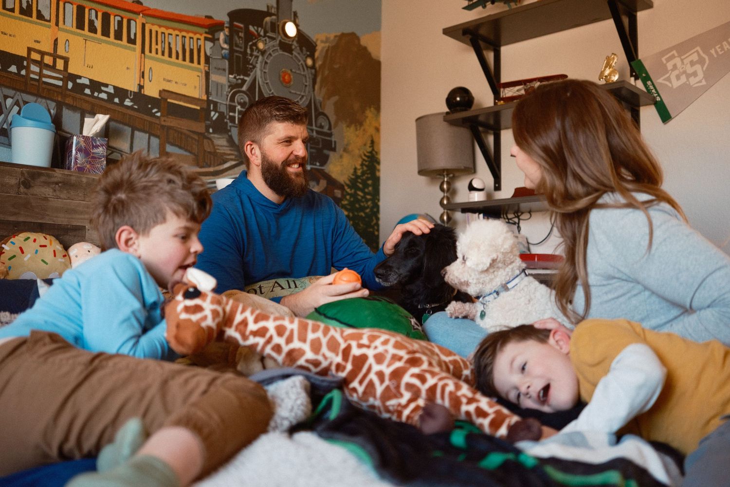 Family of two parents and two boys laughing playfully on a bed surrounded by two dogs and stuffed animals.