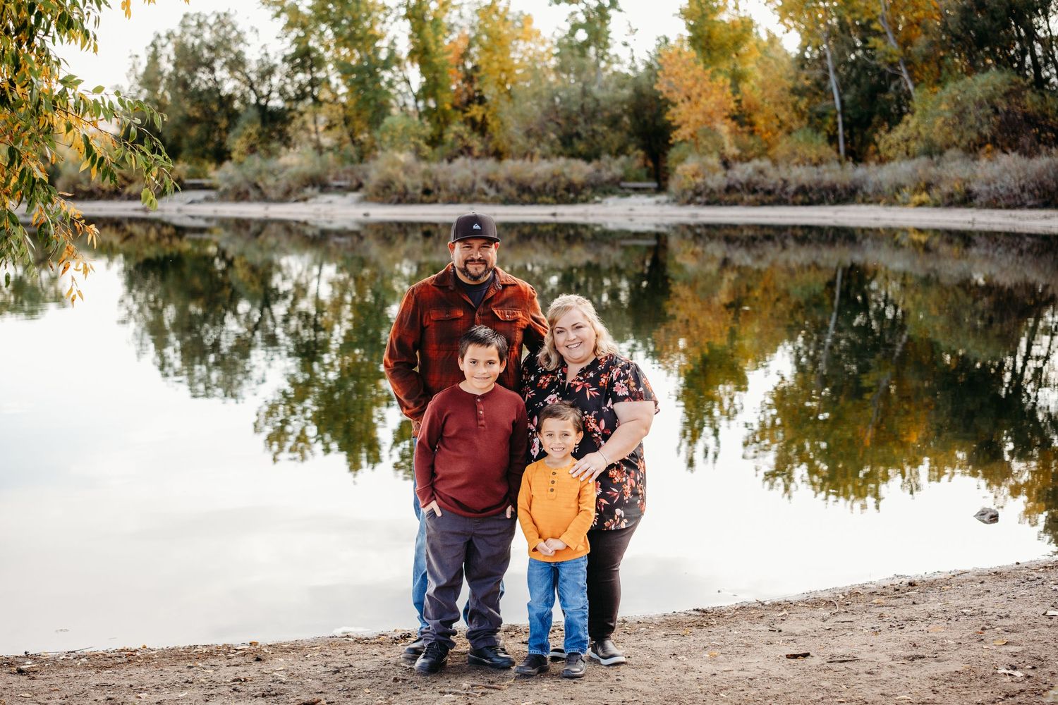 Posed family of four photo smiling at the camera, standing in front of Bear Creek Lake, framed by colorful fall trees.