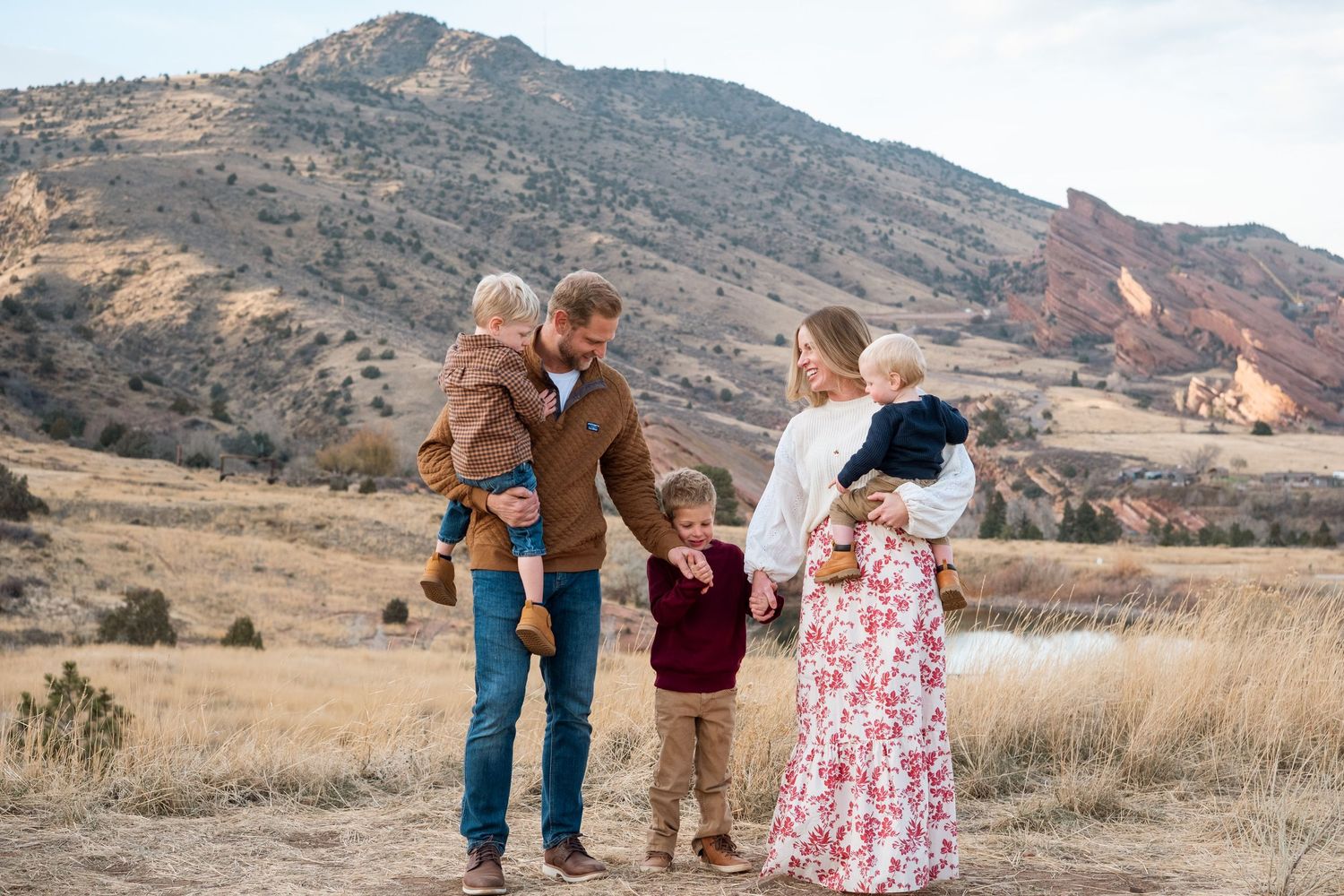Family of five standing together, holding hands and smiling at one another at Mt Falcon East in Morrison Colorado, with Red Rock views.