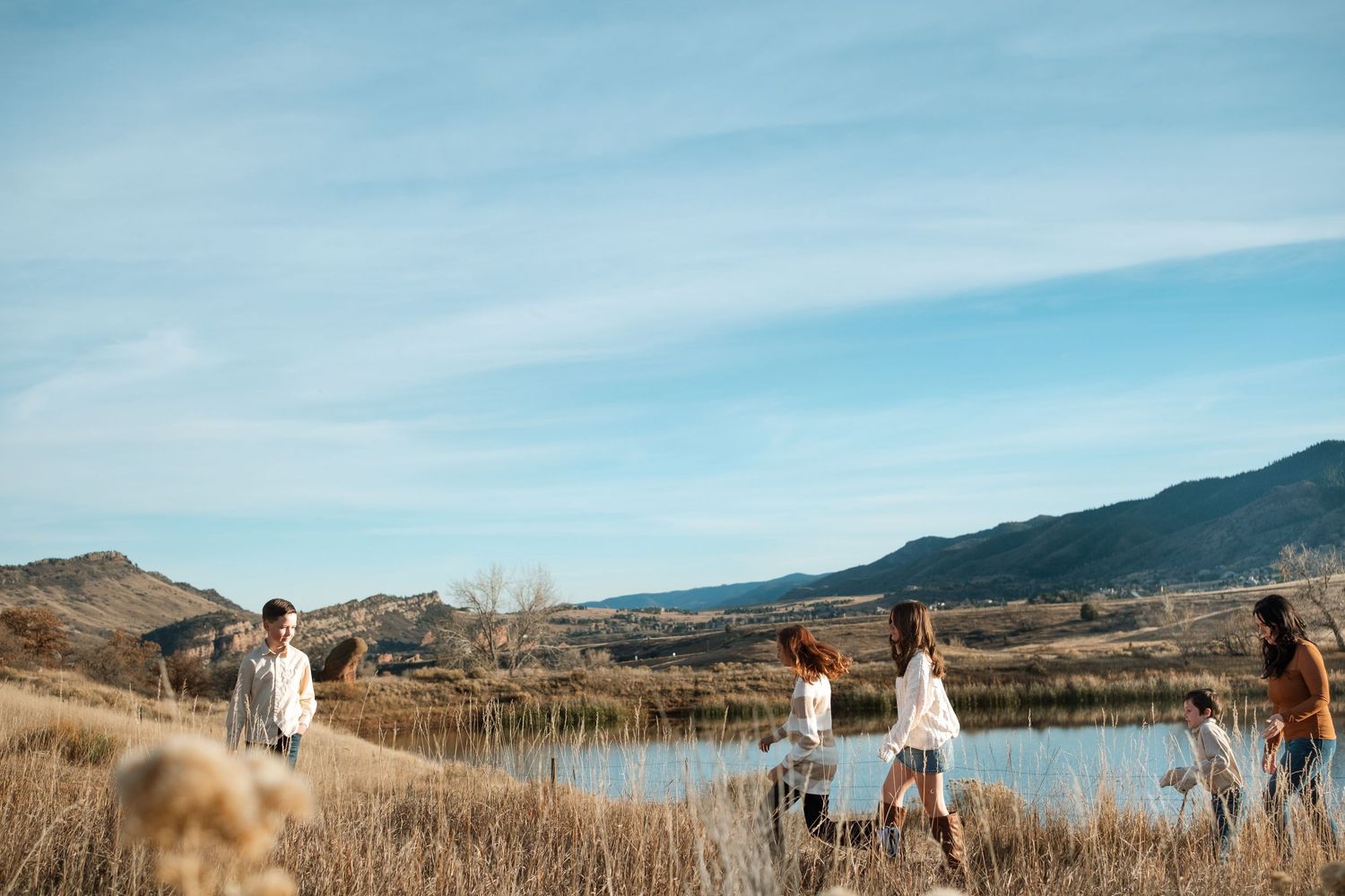 Family walking in tall grassy area, pond and foothills in the background.