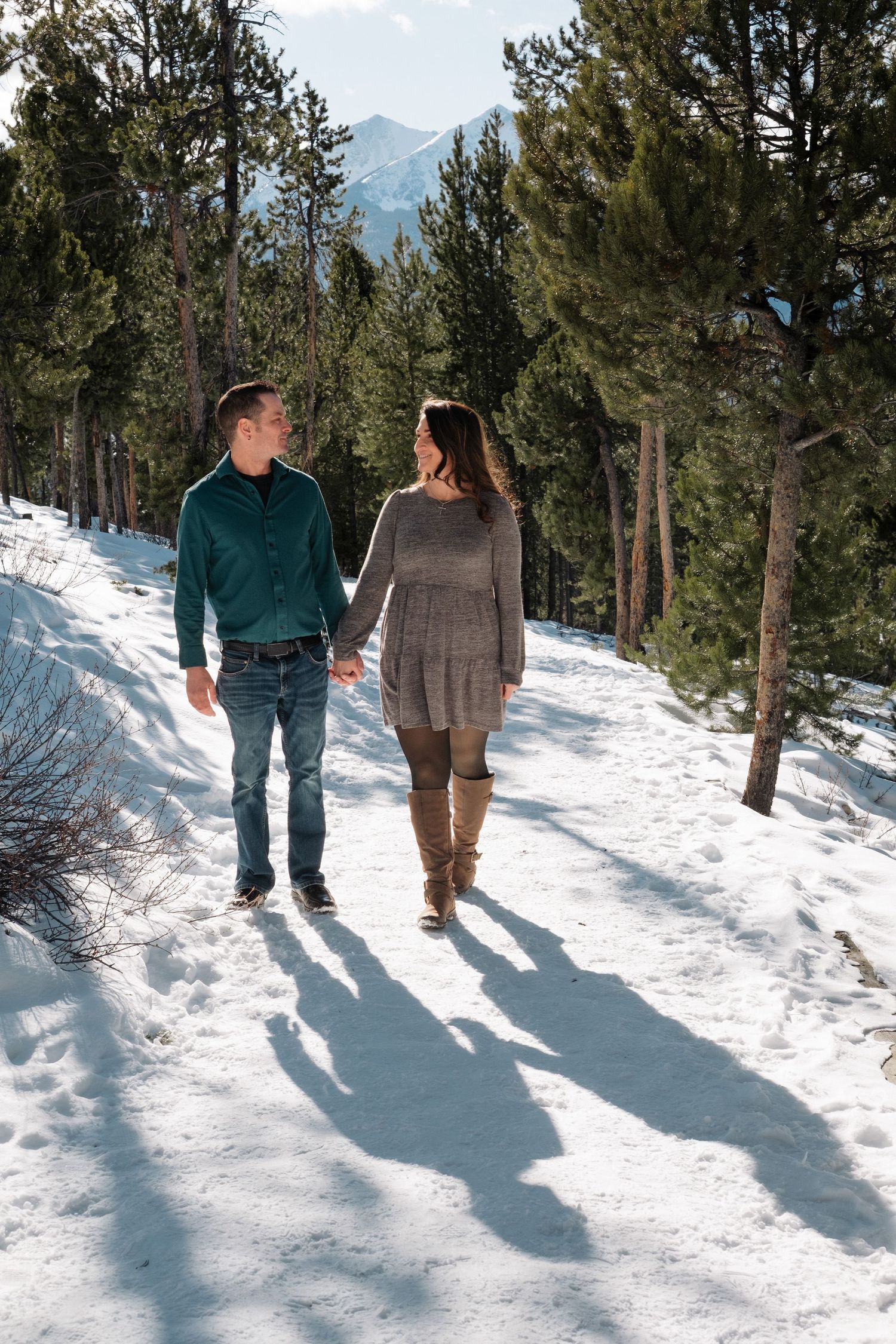 Engaged couple walking together on snowy trail at Sapphire Point Overlook, surrounded by tall trees.