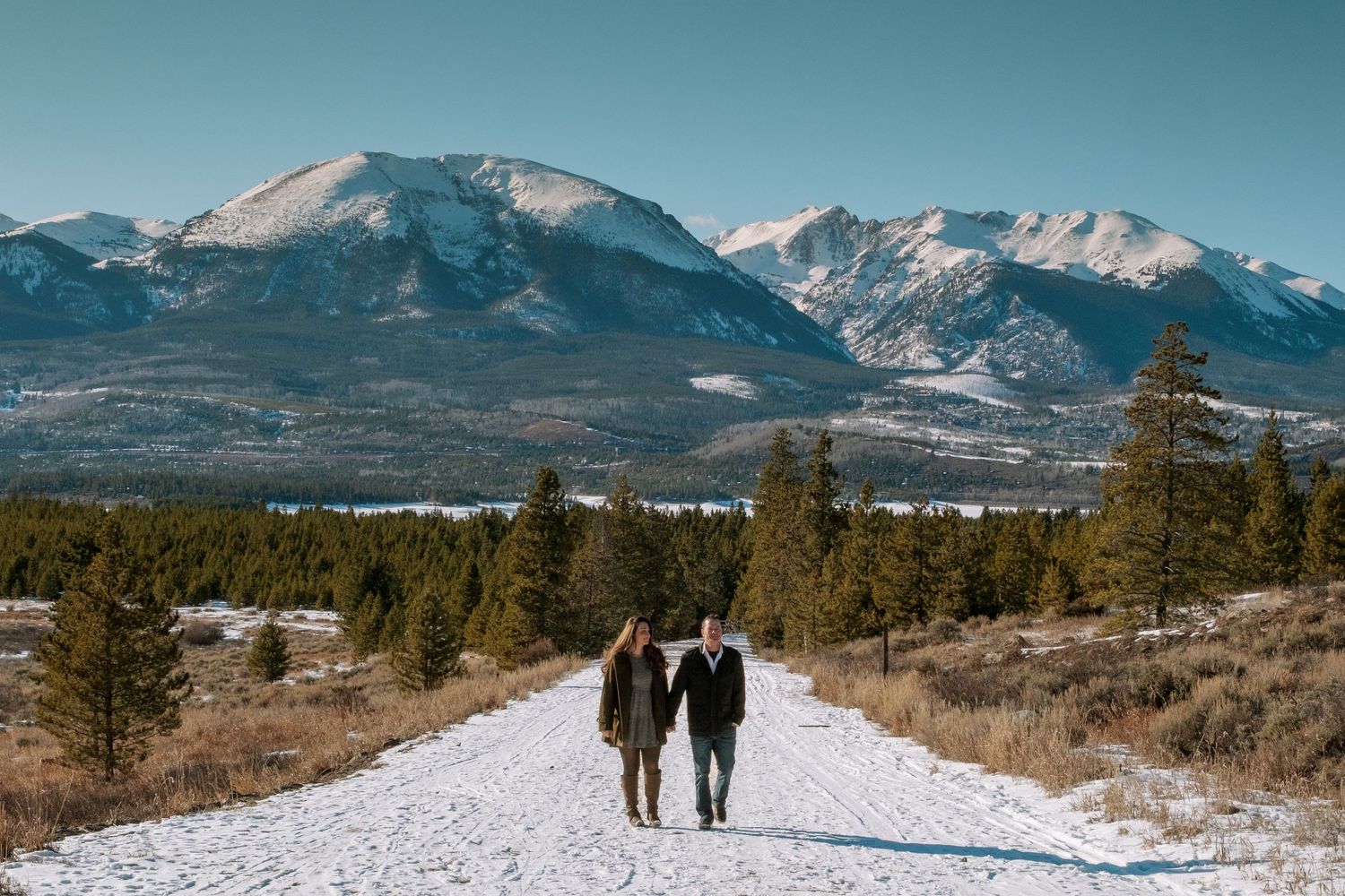 Newly engaged couple walking on snowy road, tall trees, Lake Dillon and dramatic mountains in the background at engagement photo session.