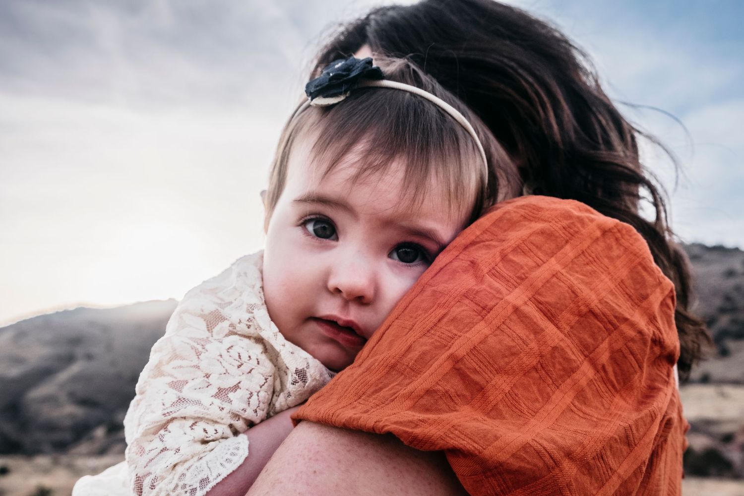 Baby girl snuggling into mom's shoulder, foothills and beautiful skies in the background.