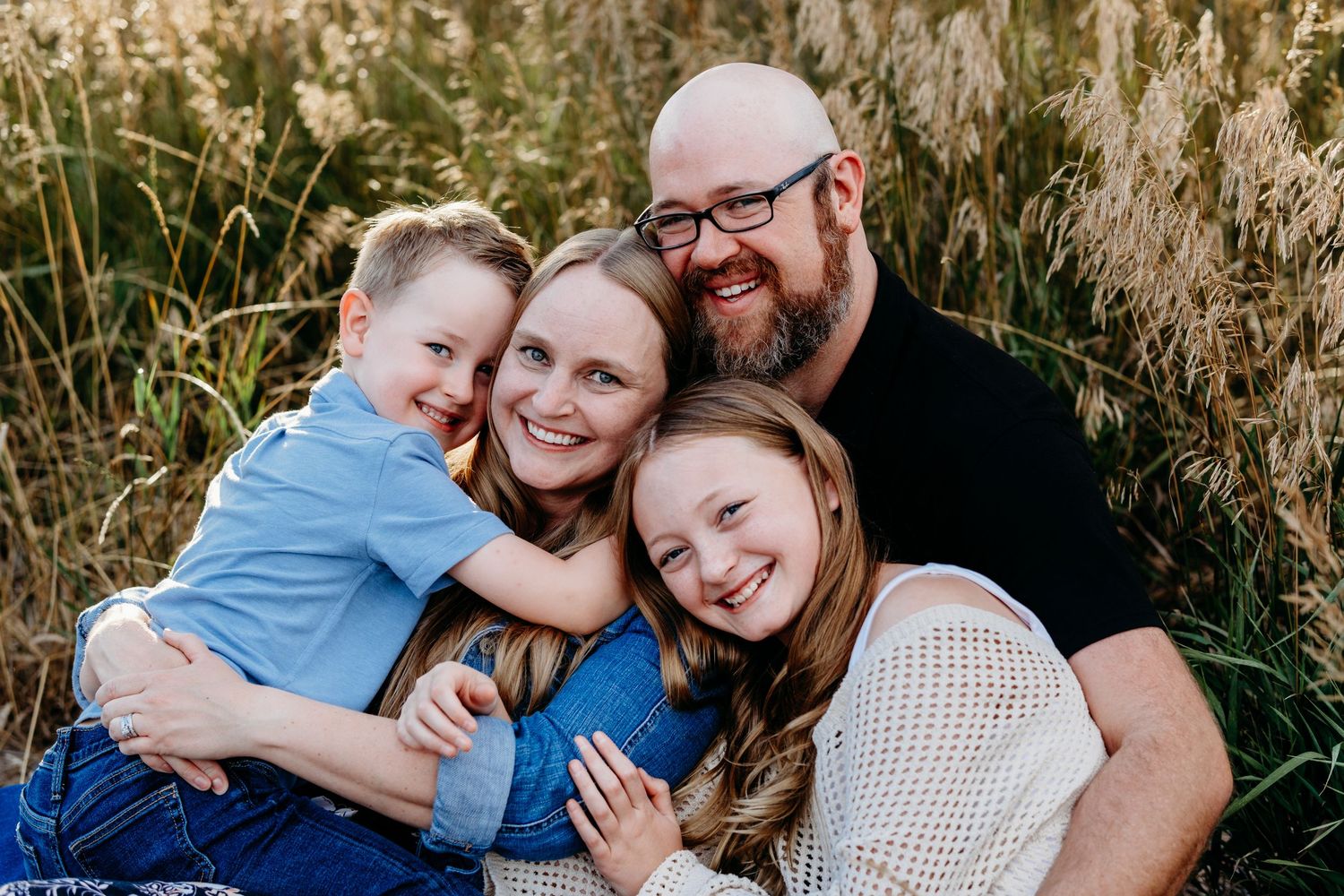 Family of father, mother, teen daughter and child son all snuggled together with warm grasses in the background.