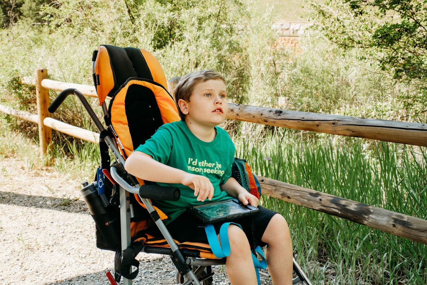 Young boy sitting in adaptive stroller with talking device on dirt path.