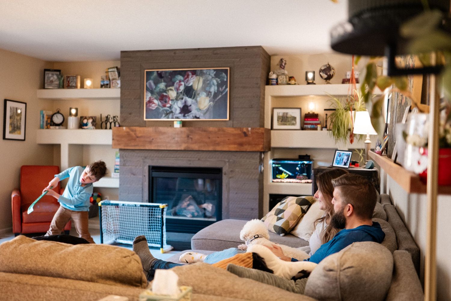 Documentary family photo of two parents sitting on the couch as son plays hockey in living room.