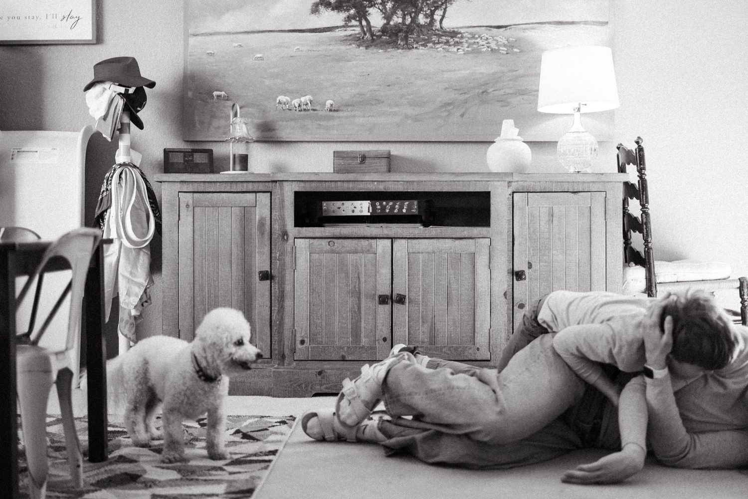 Black and white documentary photo of son wrestling with mom on the floor as a white dog looks in their direction.