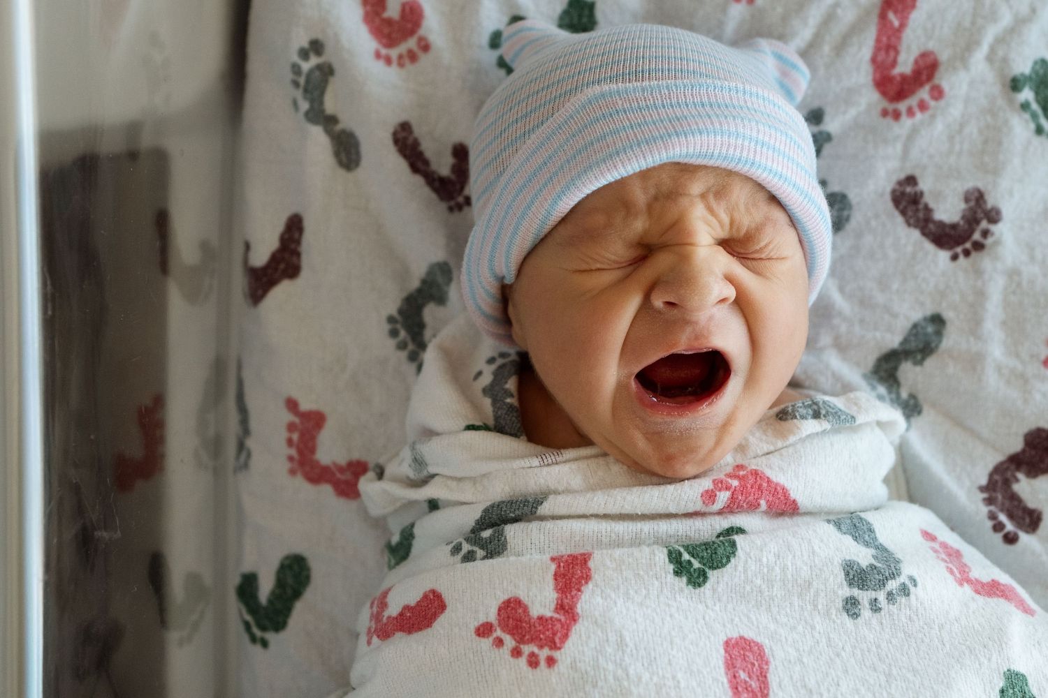 Newborn boy with hospital swaddles in hospital provided cap, newborn yawning.