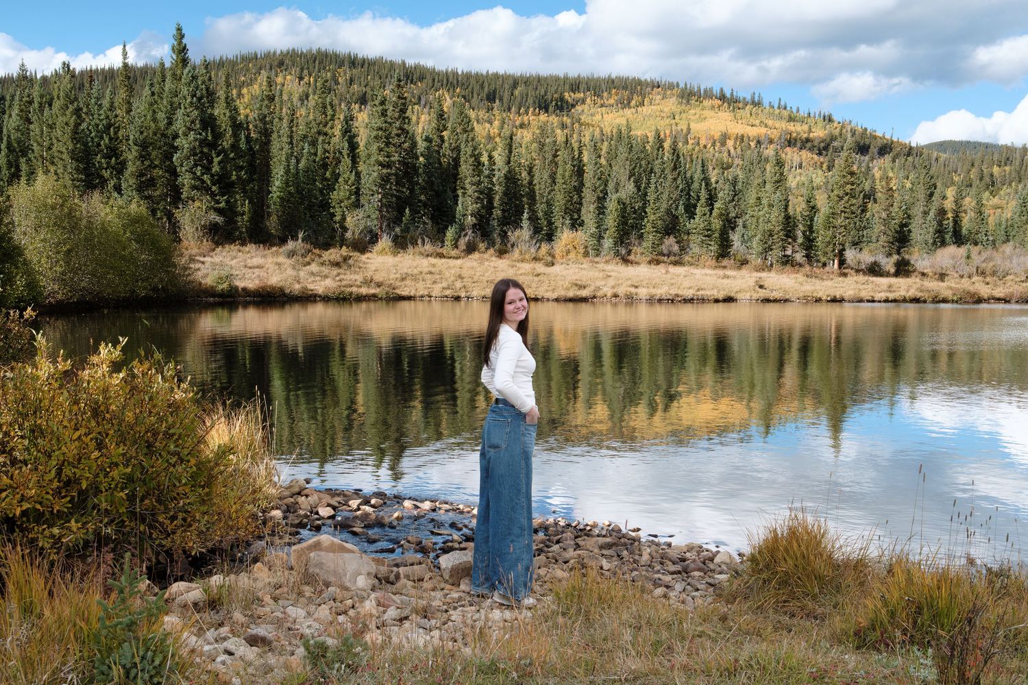Teen girl standing in front of pond in Colorado mountains with dramatic reflection in the lake.