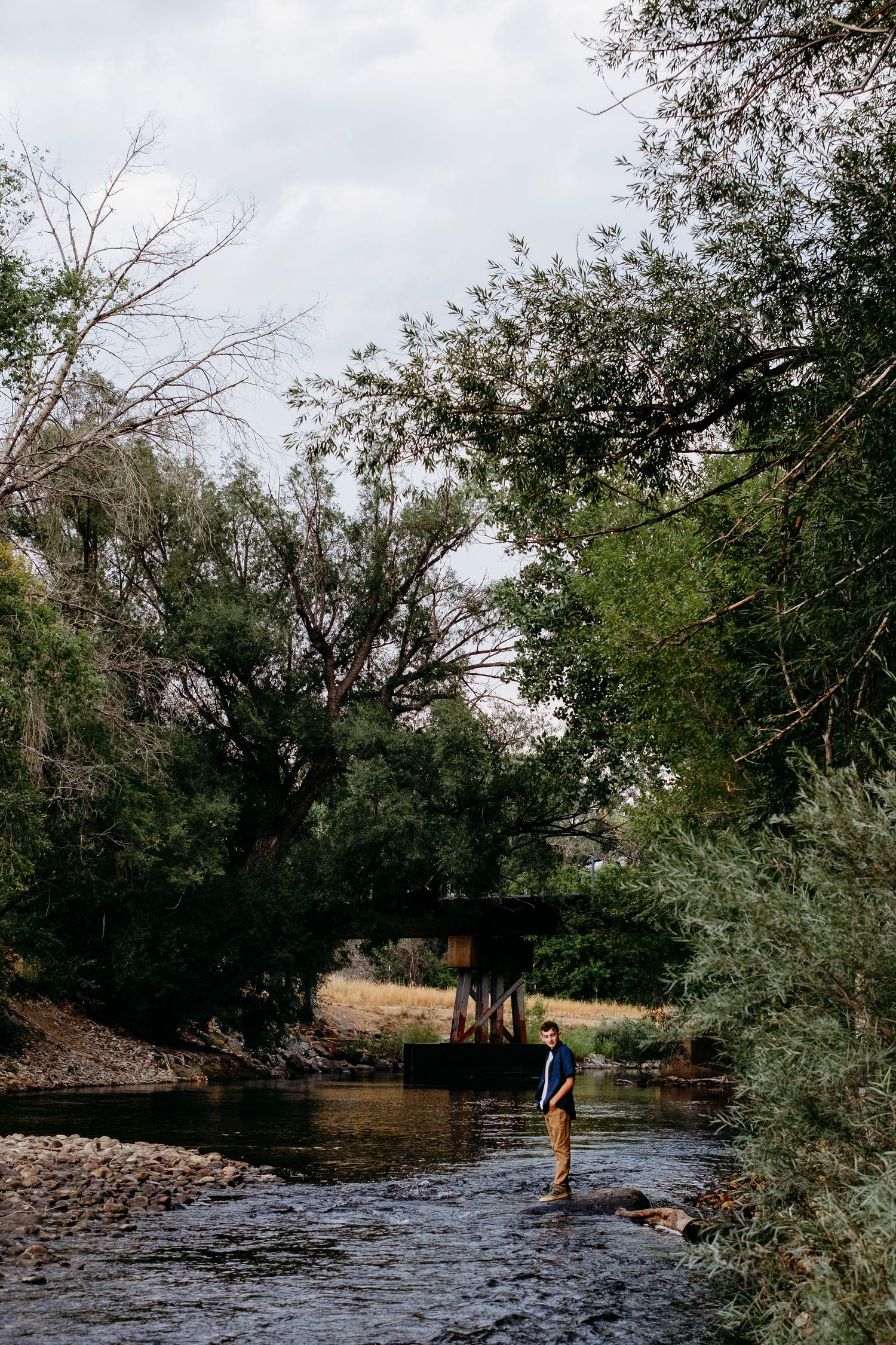 Teenage boy standing on rock off of the bank of the Cache la Poudre, ironwork bridge in the background, surrounded by lush green trees.