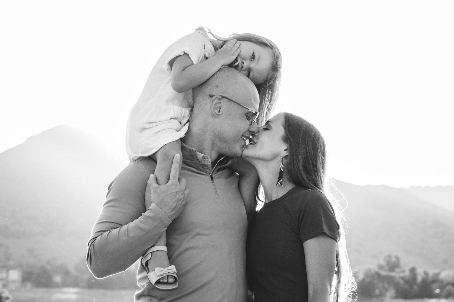 Black and white portrait of young girl perched atop father's shoulders, mother and father nearly kissing, mountains in background.